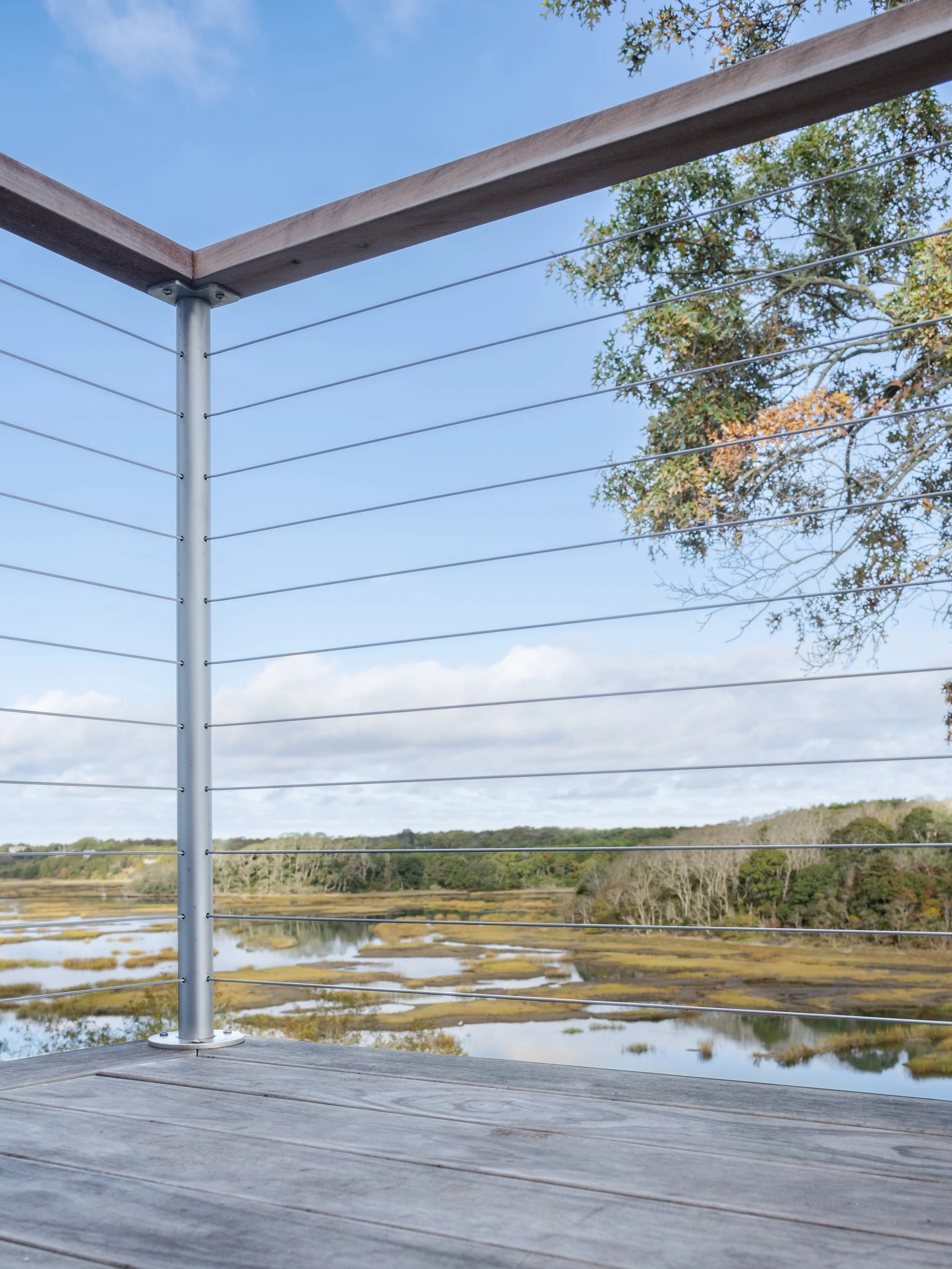 View from a wooden balcony with metal railing overlooking a marshland with water, trees, and a partly cloudy blue sky.