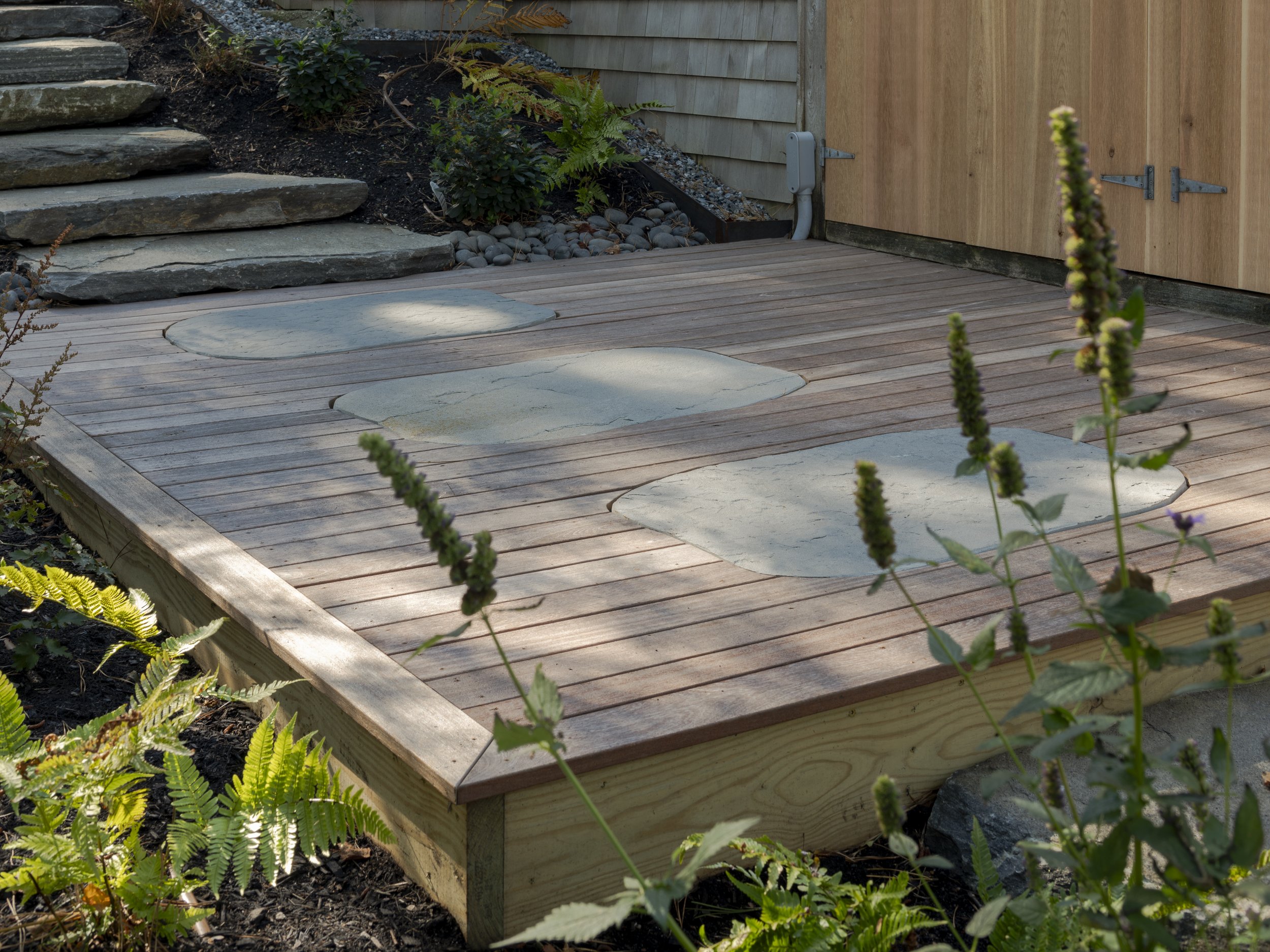 A wooden deck with three embedded stone steps, surrounded by plants and a garden bed with rocks, and a wooden shed door.
