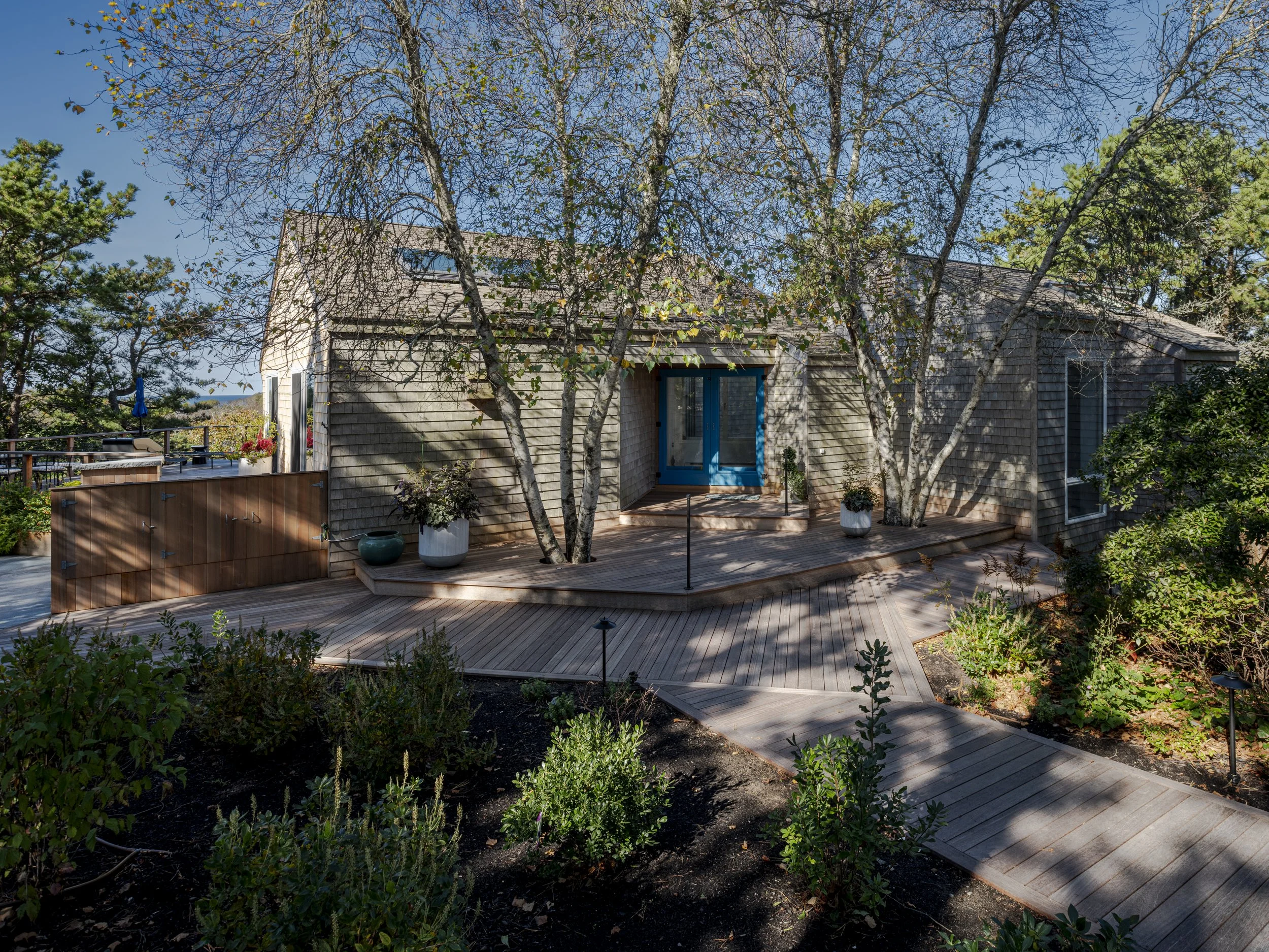 A house with gray shingle siding, blue front door, and a wooden deck with potted plants, surrounded by trees and a garden.