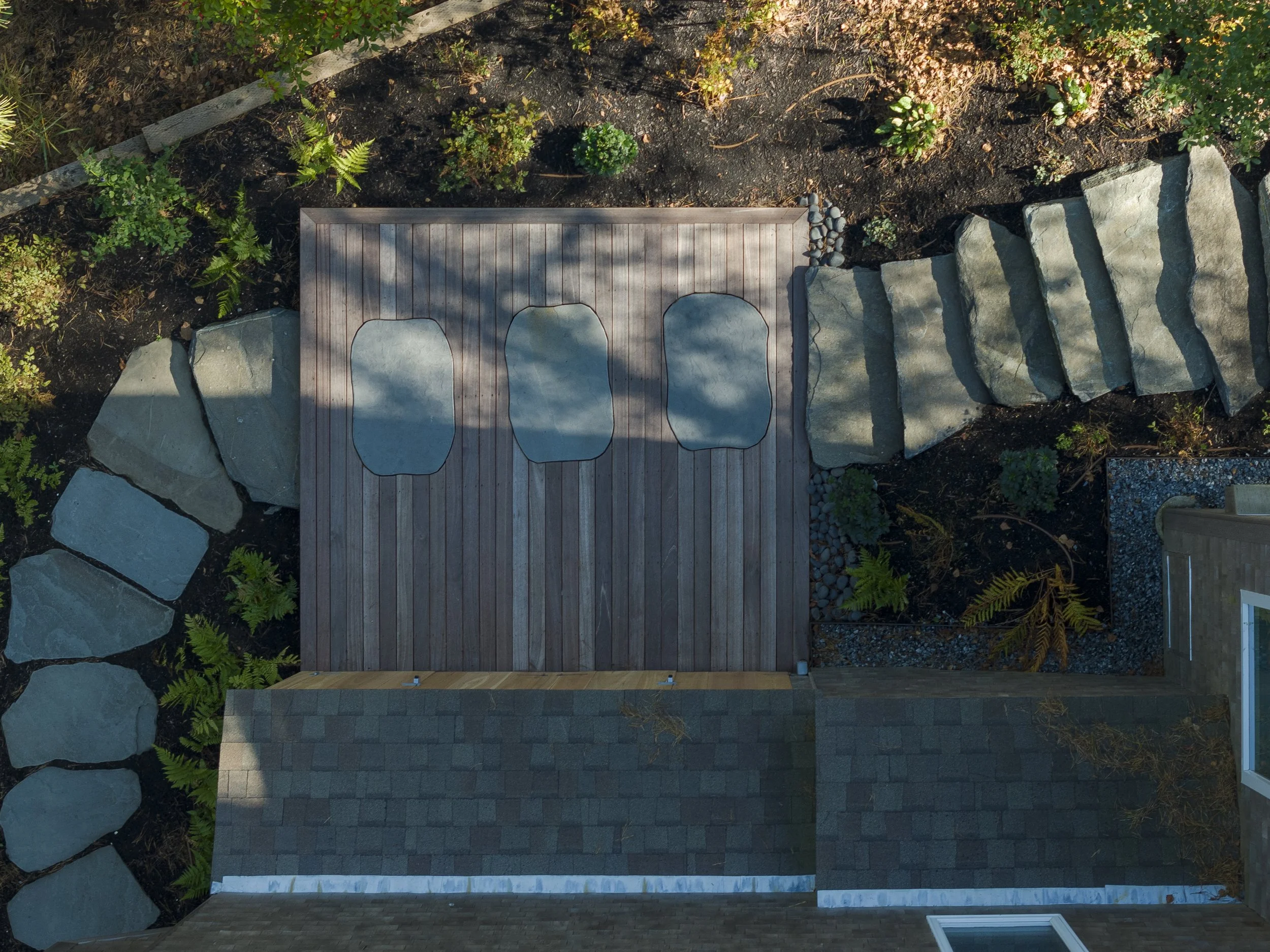 Overhead view of a backyard patio with three round non-woven mats on a wooden deck, stone stepping stones bordering a flower bed, arranged on dark soil with various green plants and foliage.