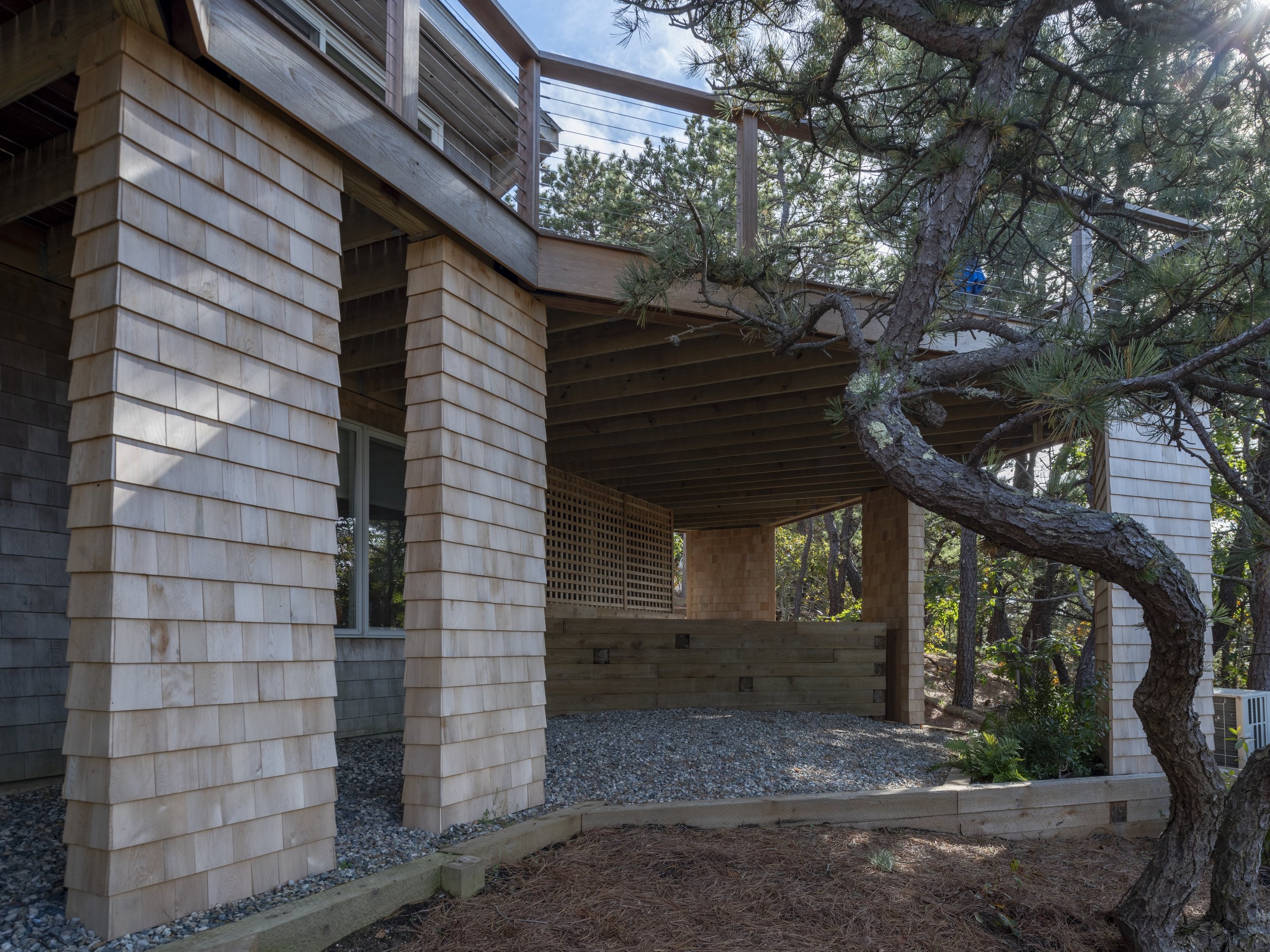 View of a wooden house with a covered porch supported by large, angled wooden columns, and a tree with twisted branches in the foreground, surrounded by trees and natural landscaping.