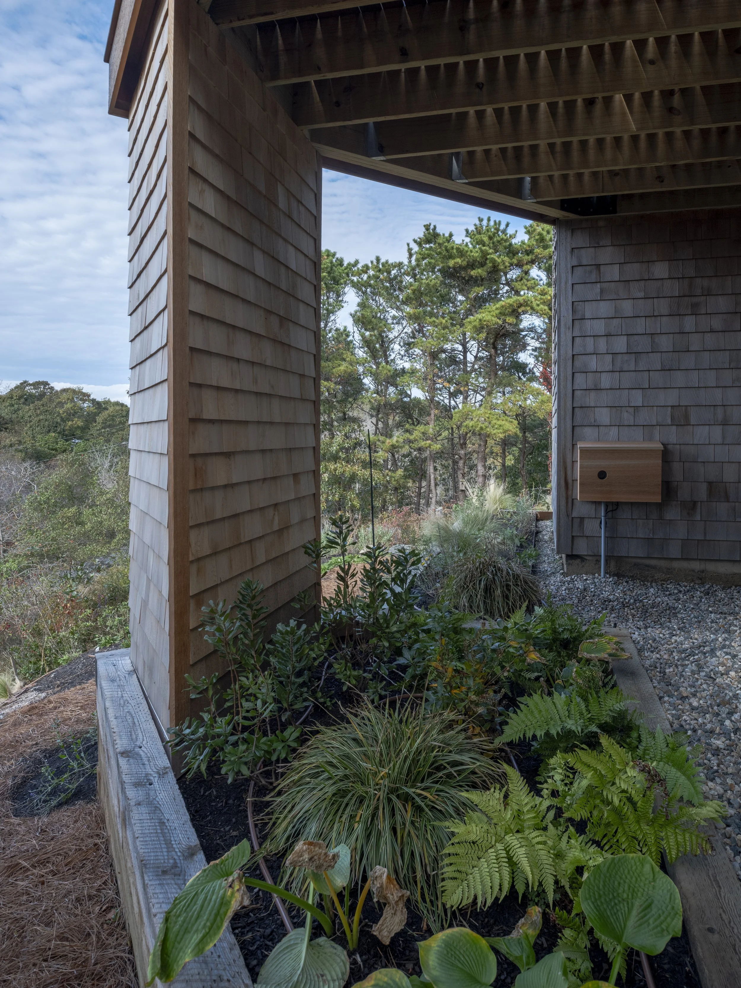 View of a wooden house corner with a garden bed, plants, and trees in the background, under a cloudy sky.