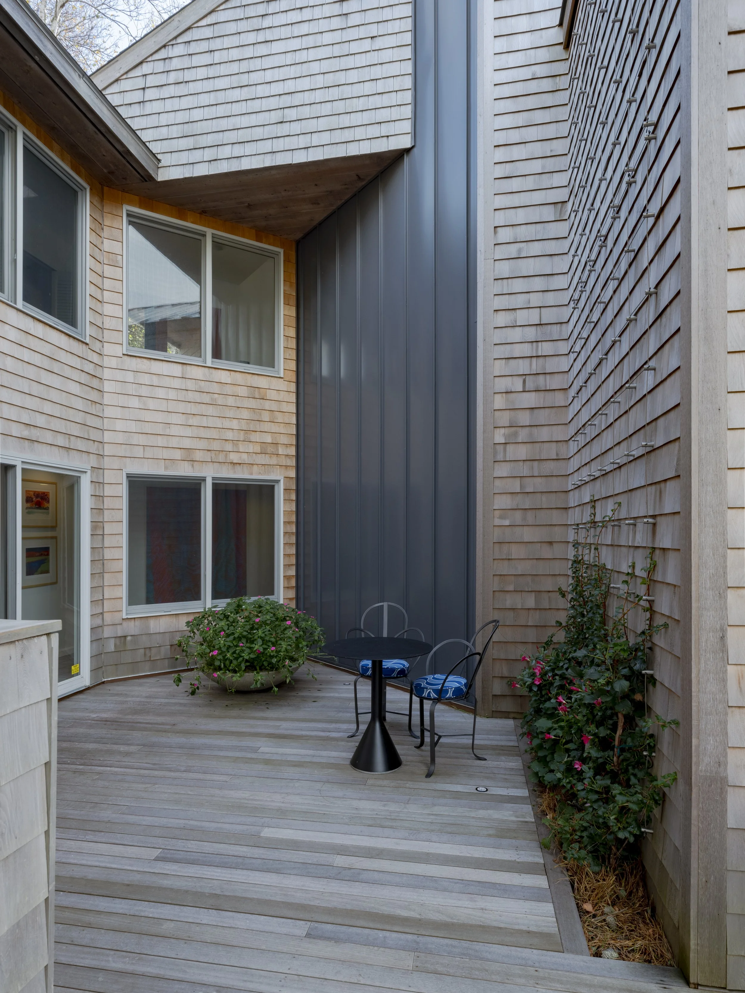 Small outdoor patio with a metal table, two chairs with blue cushions, potted plants, and wooden and metal exterior walls of a building.