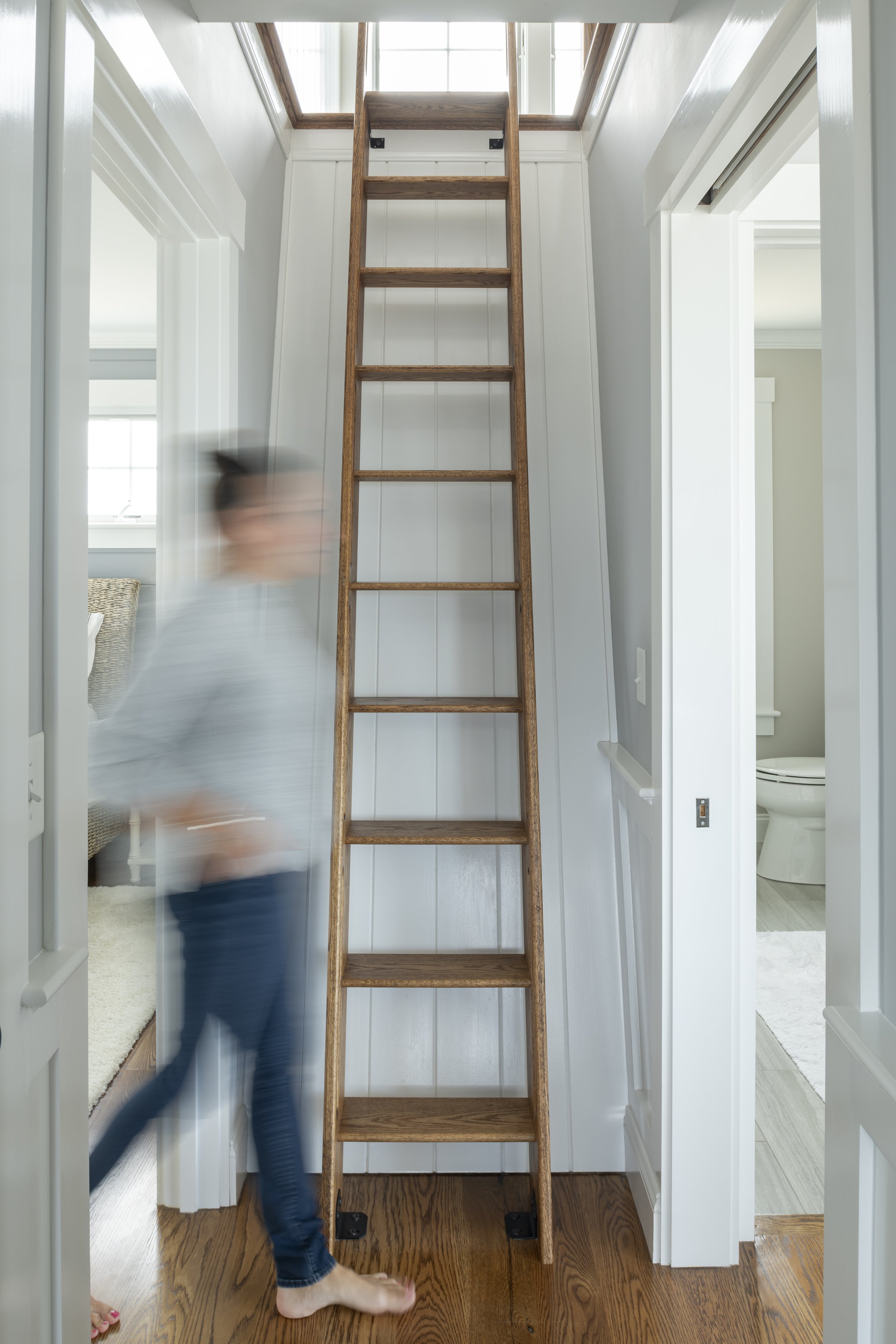 A wooden ladder leading up to a skylight window in the ceiling of a bright room; a person is blurred, walking past in the foreground.