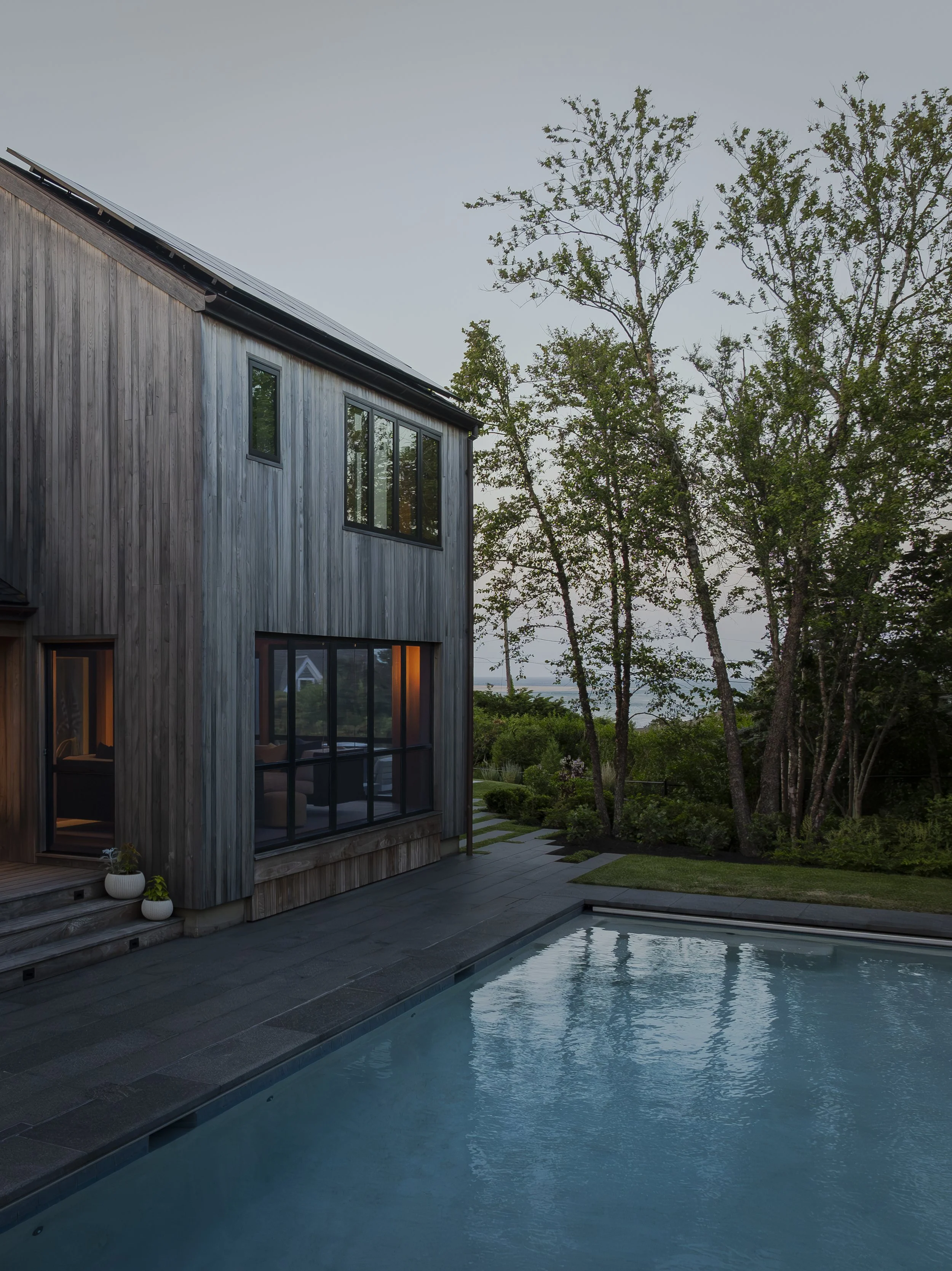 A modern house with large windows and wooden exterior, overlooking a swimming pool with water reflecting the sky, surrounded by trees and greenery at dusk.