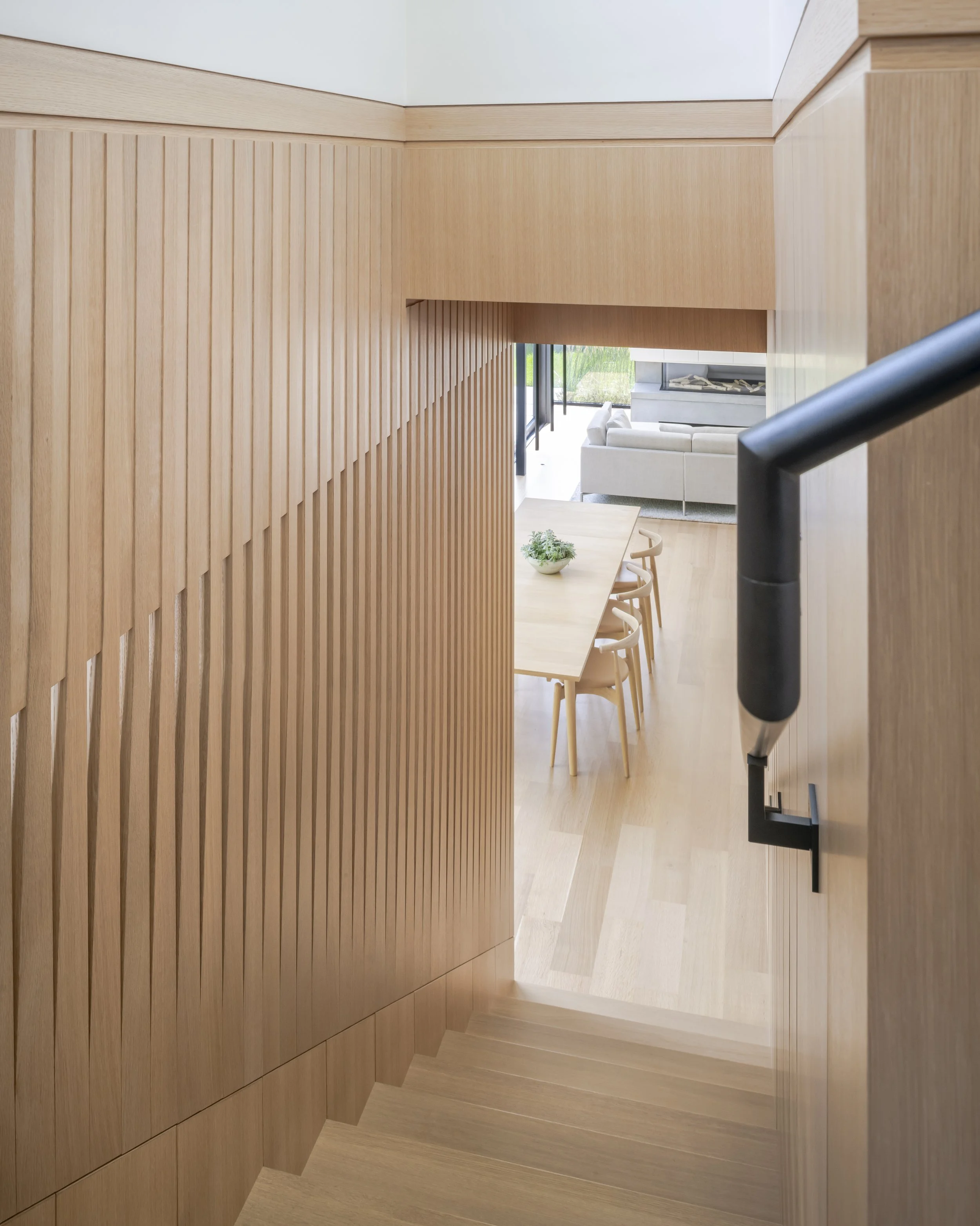 Wood-paneled staircase leading to a dining and living area with a wooden dining table, chairs, a white sofa, and a fireplace.