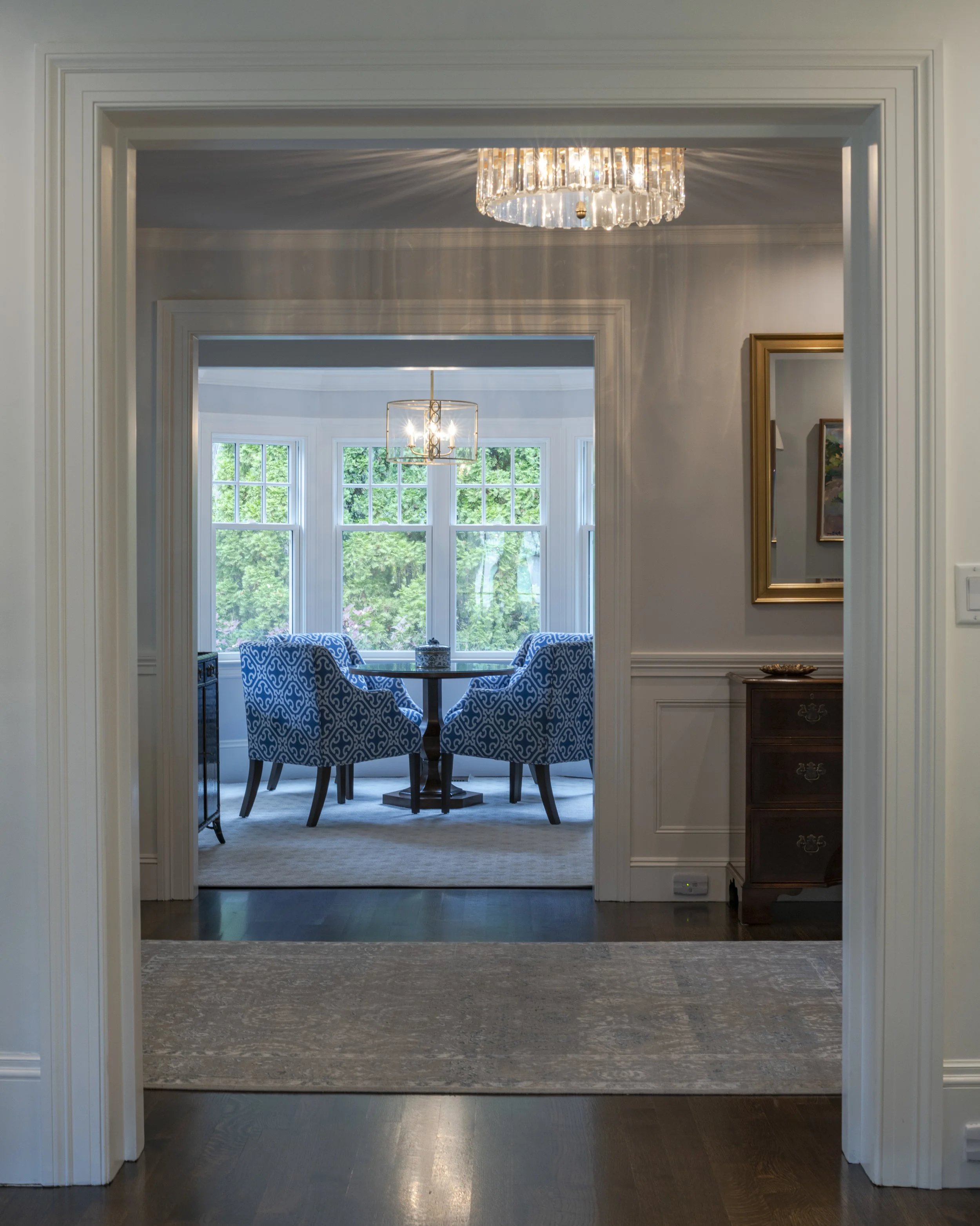 View through a doorway into a dining area with four blue patterned armchairs around a dark wooden table near a window with green trees outside. A modern chandelier hangs from the ceiling.