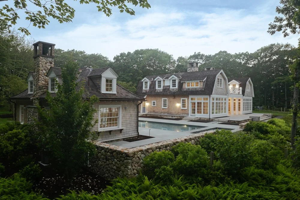 A large, luxurious house with multiple stories and a stone chimney, surrounded by lush greenery, with a swimming pool in the backyard.