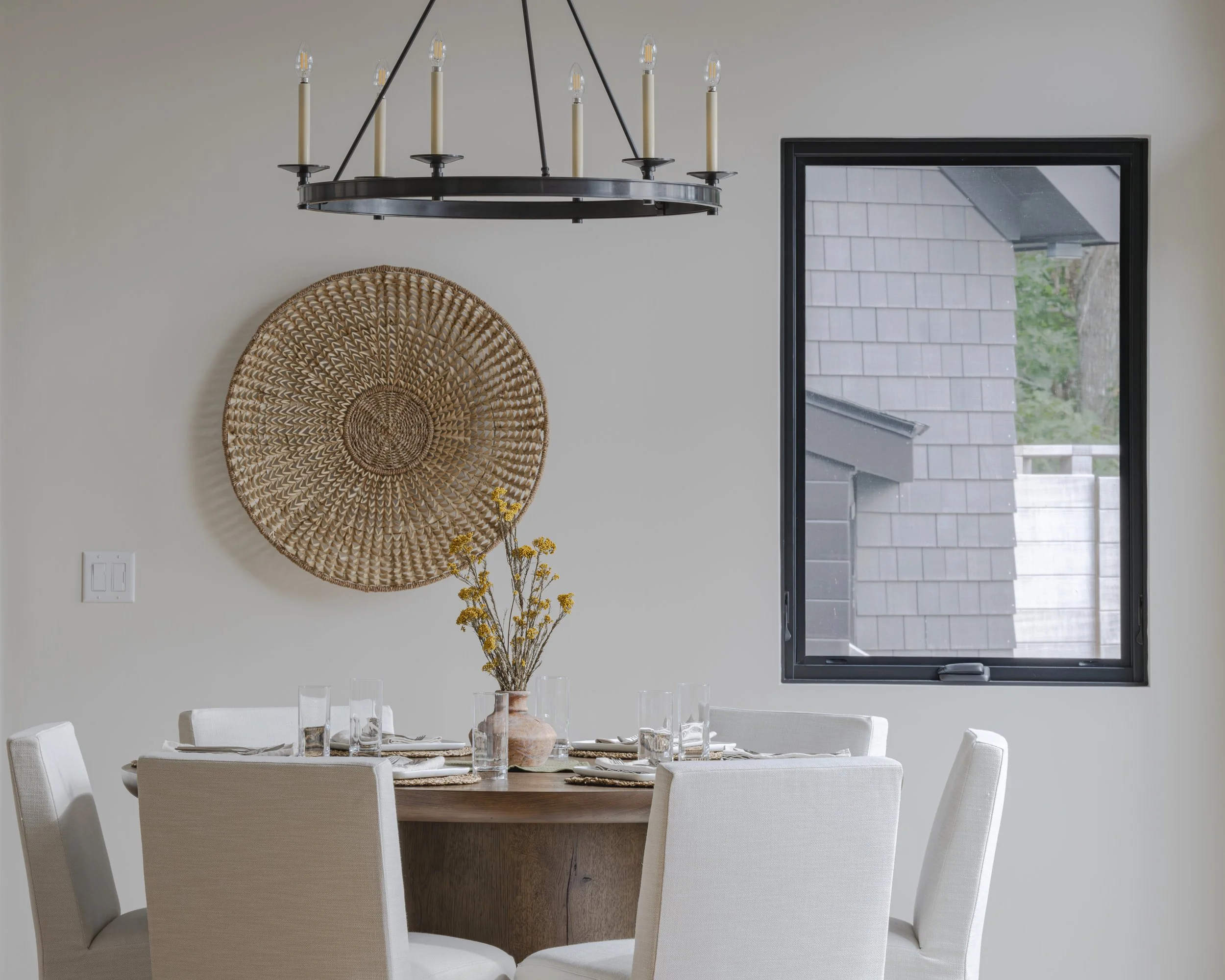 A dining room with a round wooden table surrounded by six white upholstered chairs, a ceramic vase with yellow flowers on the table, a large woven wall art piece, a black chandelier hanging above, and a window showing a neighboring house's roof.
