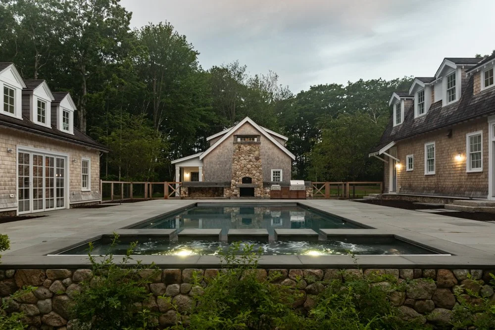 A backyard with a rectangular swimming pool surrounded by concrete paving, two houses with shingle siding and dormer windows on either side, a stone and wood fireplace or grill structure at the far end, and lush green trees behind fences or railing.