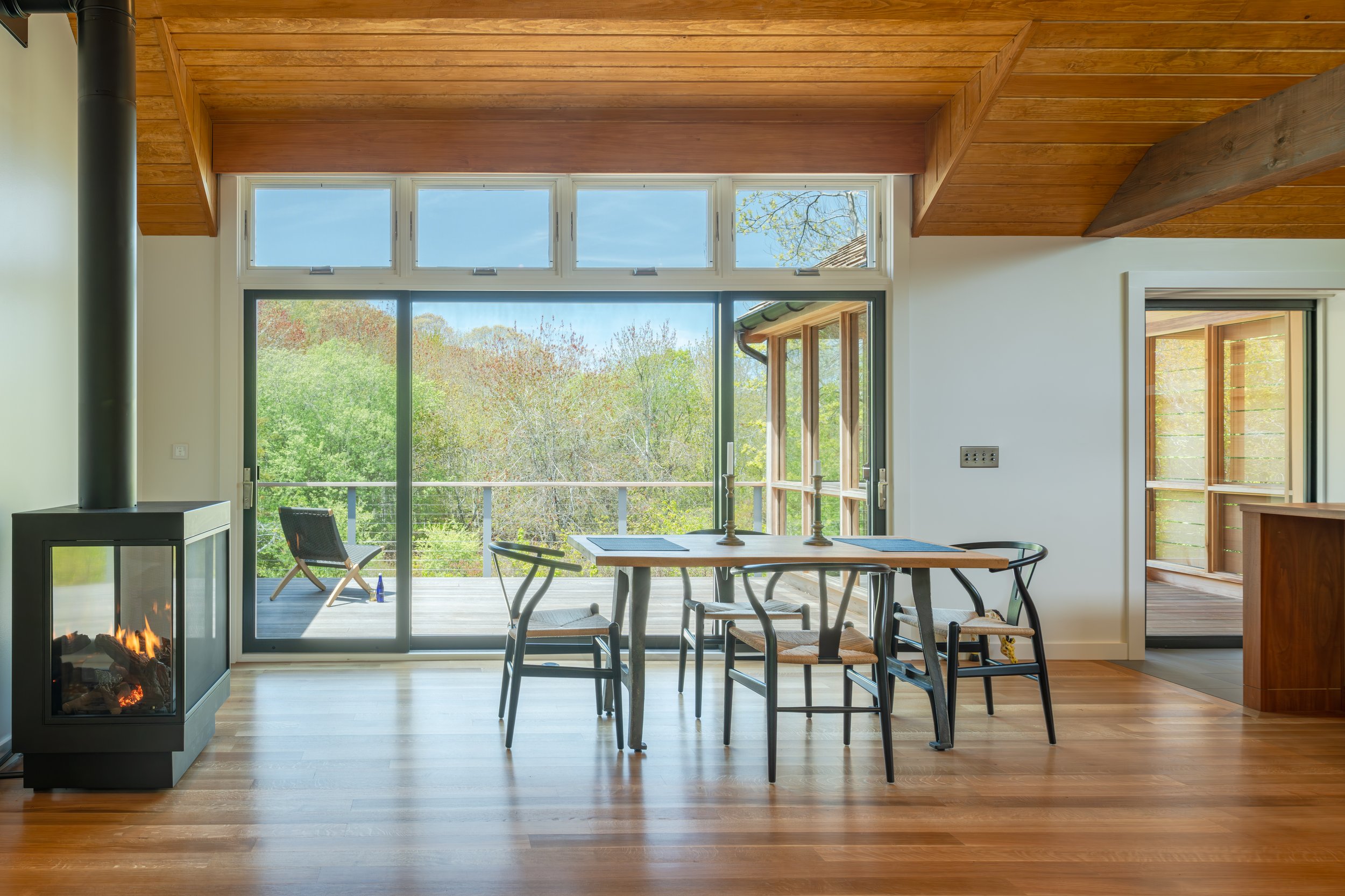 Bright dining area with a wooden ceiling, large glass sliding door and window, a wooden table with four black chairs, a small fireplace with a burning fire, and a balcony with outdoor seating overlooking trees.