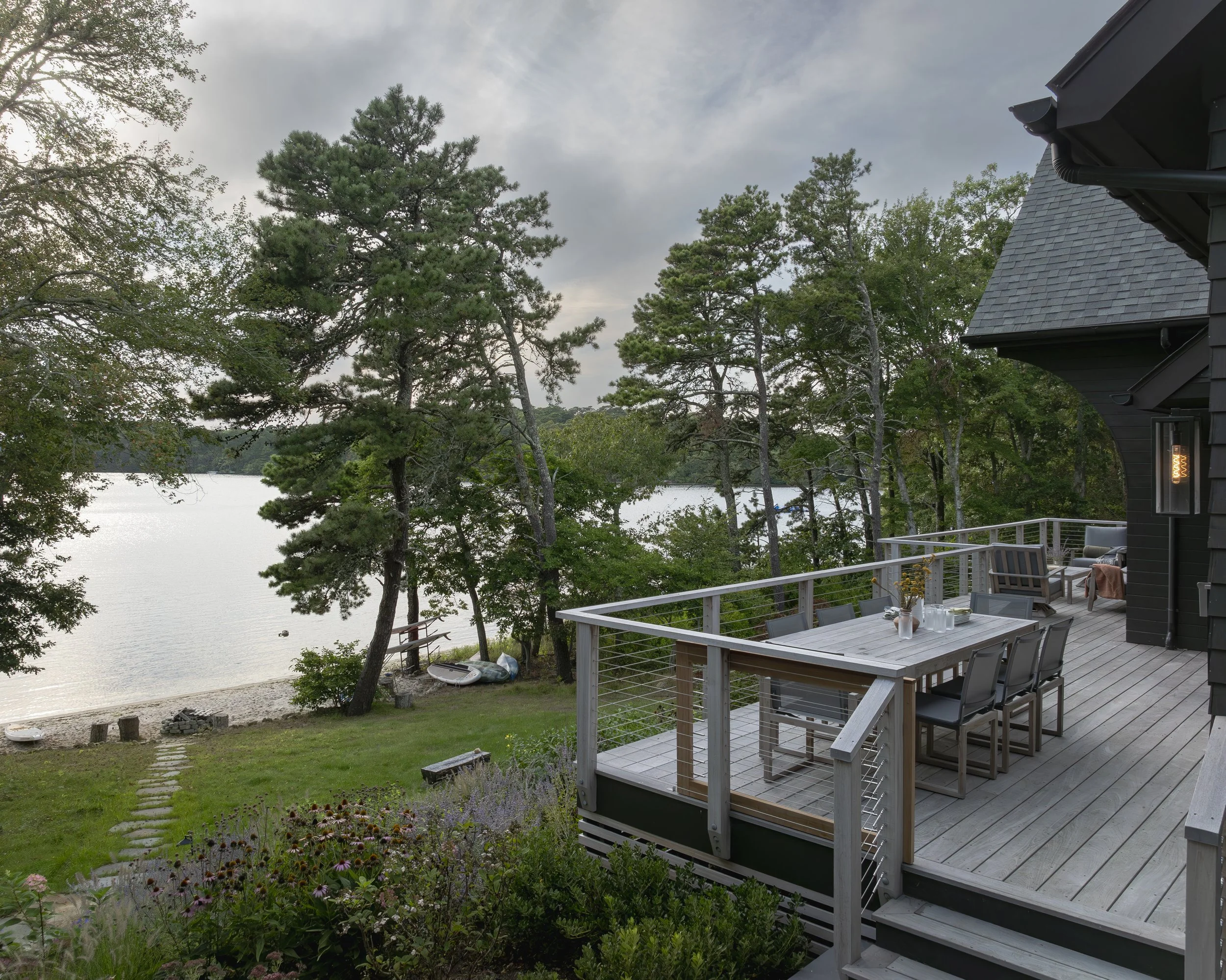 A house with a large outdoor deck overlooking a lake, surrounded by trees. The deck has a dining table with chairs, a bench, and a seating area with cushions. There is a pathway leading to the lake and a small boat on the shore.