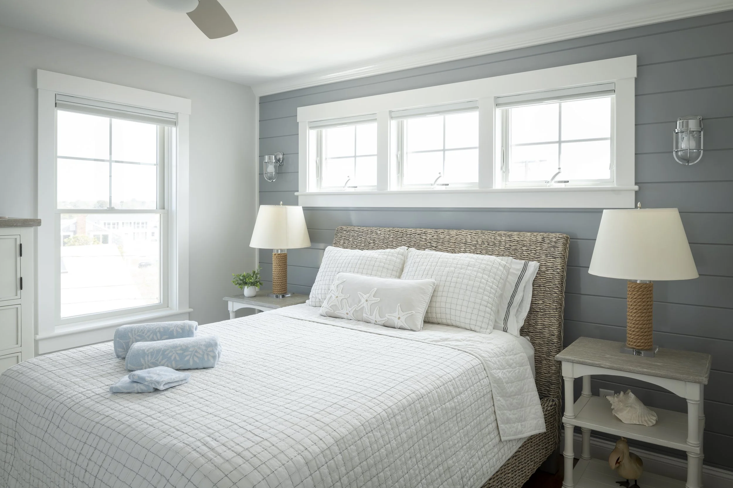 Bright bedroom with a white bed, wicker headboard, and nautical decor, featuring two white nightstands with lamps, on a gray paneled wall with three windows.