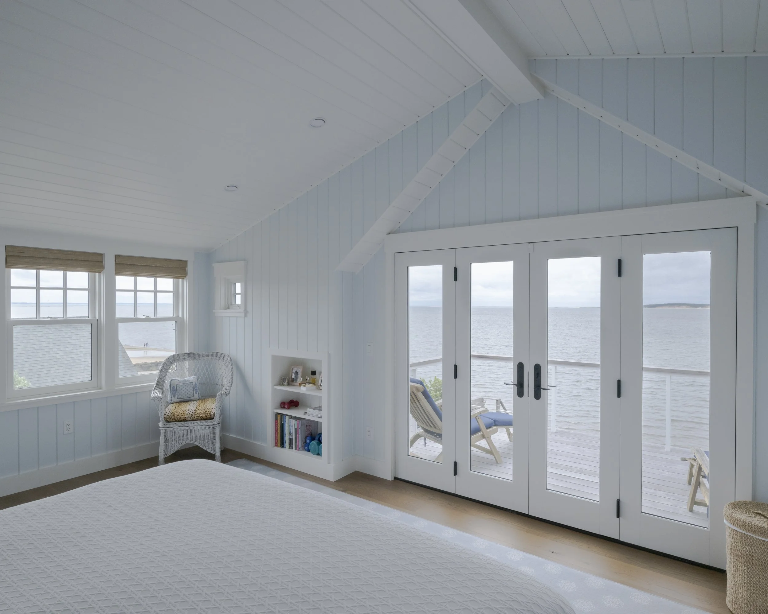 Cozy bedroom with ocean view, featuring white walls, a large bed, a wicker chair with cushions, and glass double doors leading to a balcony facing the water.