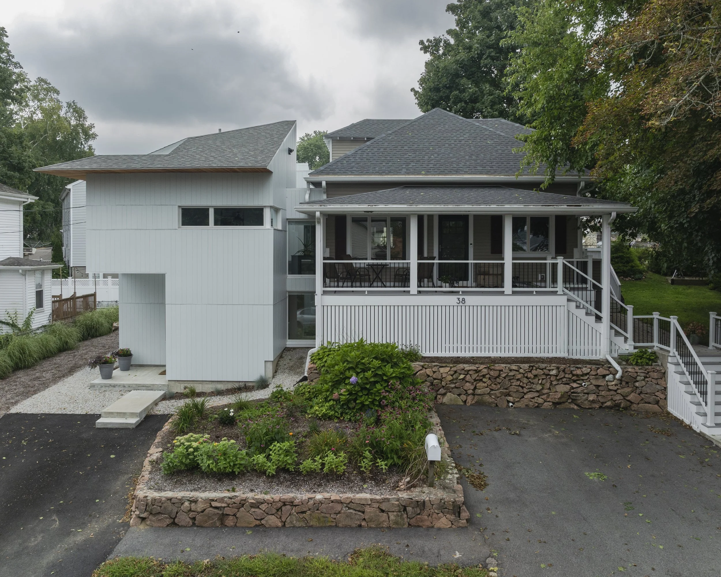 A two-story house combines modern and traditional design elements, with a gray traditional roof, a front porch with railing, and white siding on the exterior. The house has a visible house number 38, a landscaped front yard with a flower bed, a drive
