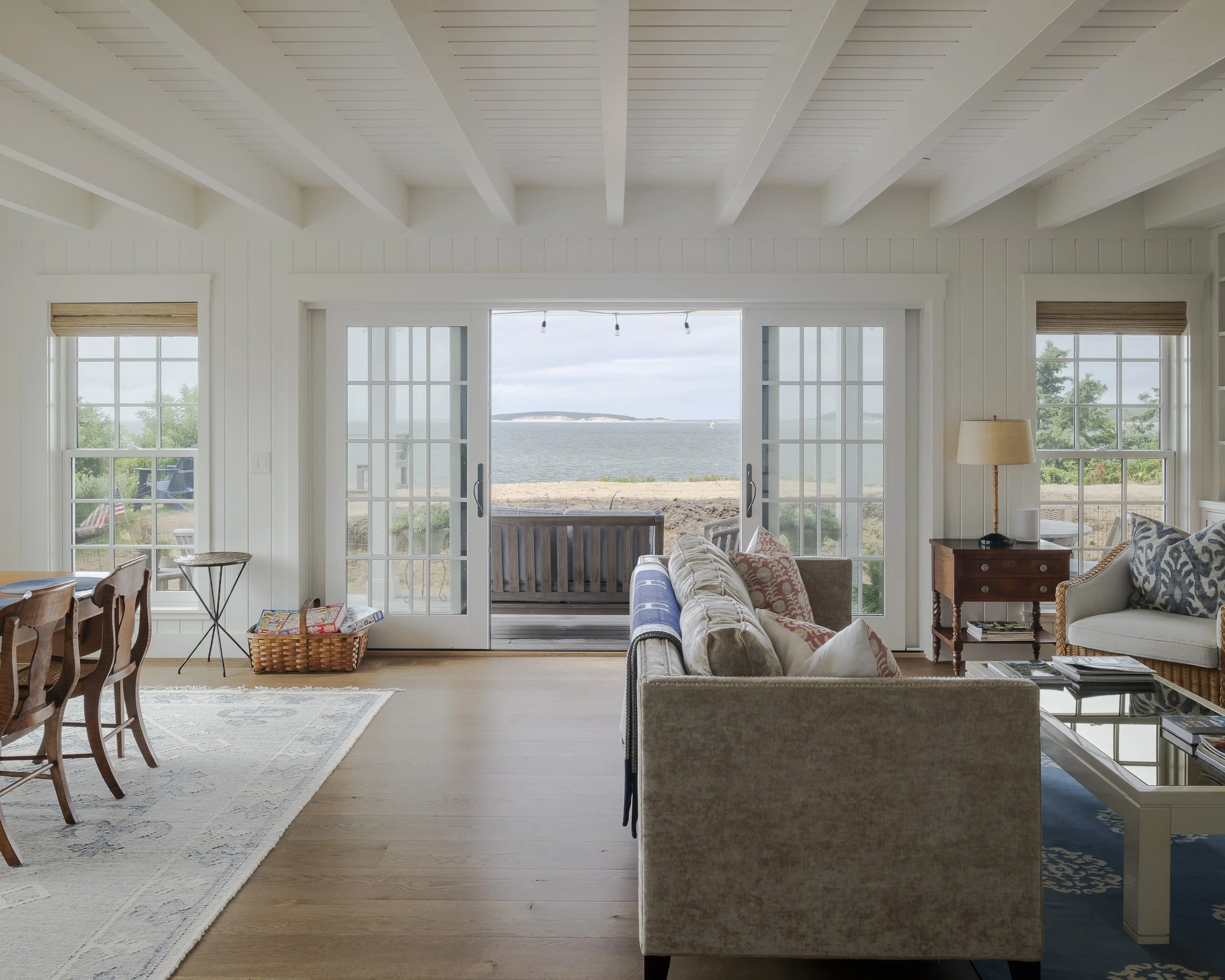 Living room with large sliding glass doors opening to a beach view with sand, water, and distant land. Interior includes a beige sofa, side table with lamp, armchair, coffee table, and various pillows. Wooden floors and windows with blinds.