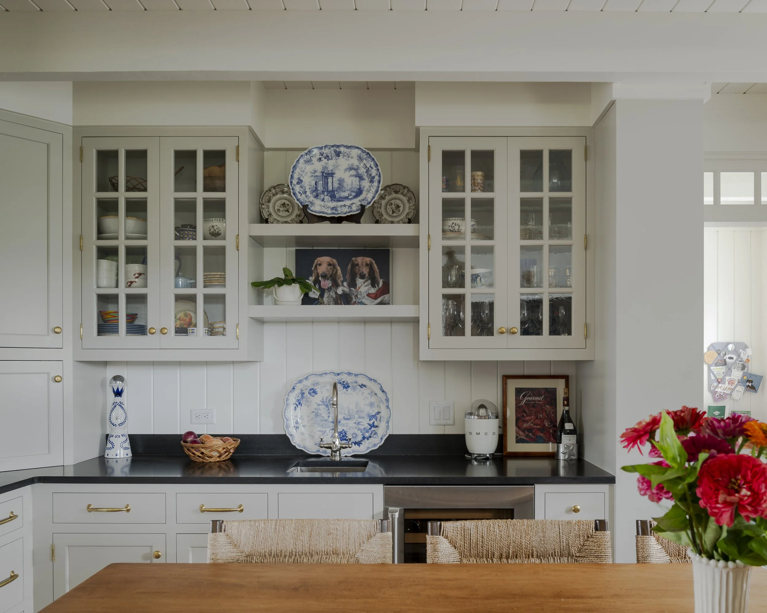 A kitchen with white cabinets, black countertops, and decorative blue and white plates and dishes displayed on shelves. There is a framed photo of two dogs, a small plant, and various kitchen items on the counter. A vase of pink and red flowers is on