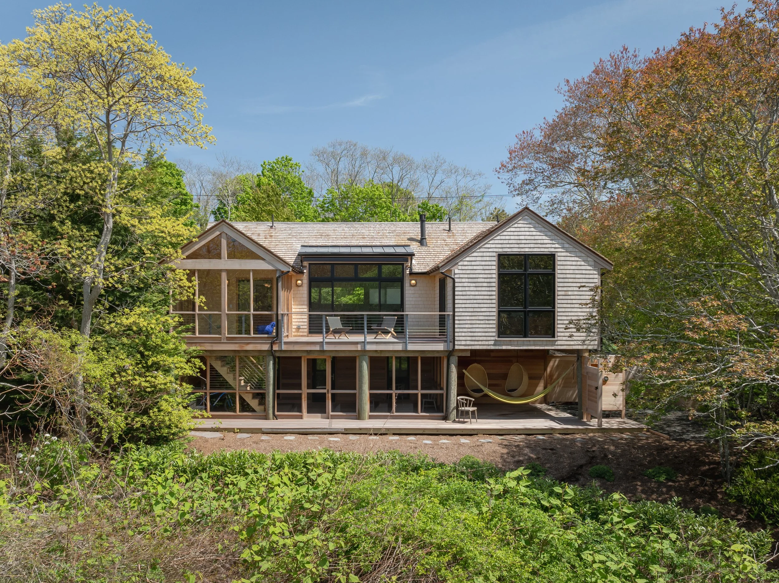 A modern two-story house with a wooden exterior, large windows, and a balcony surrounded by trees and greenery.