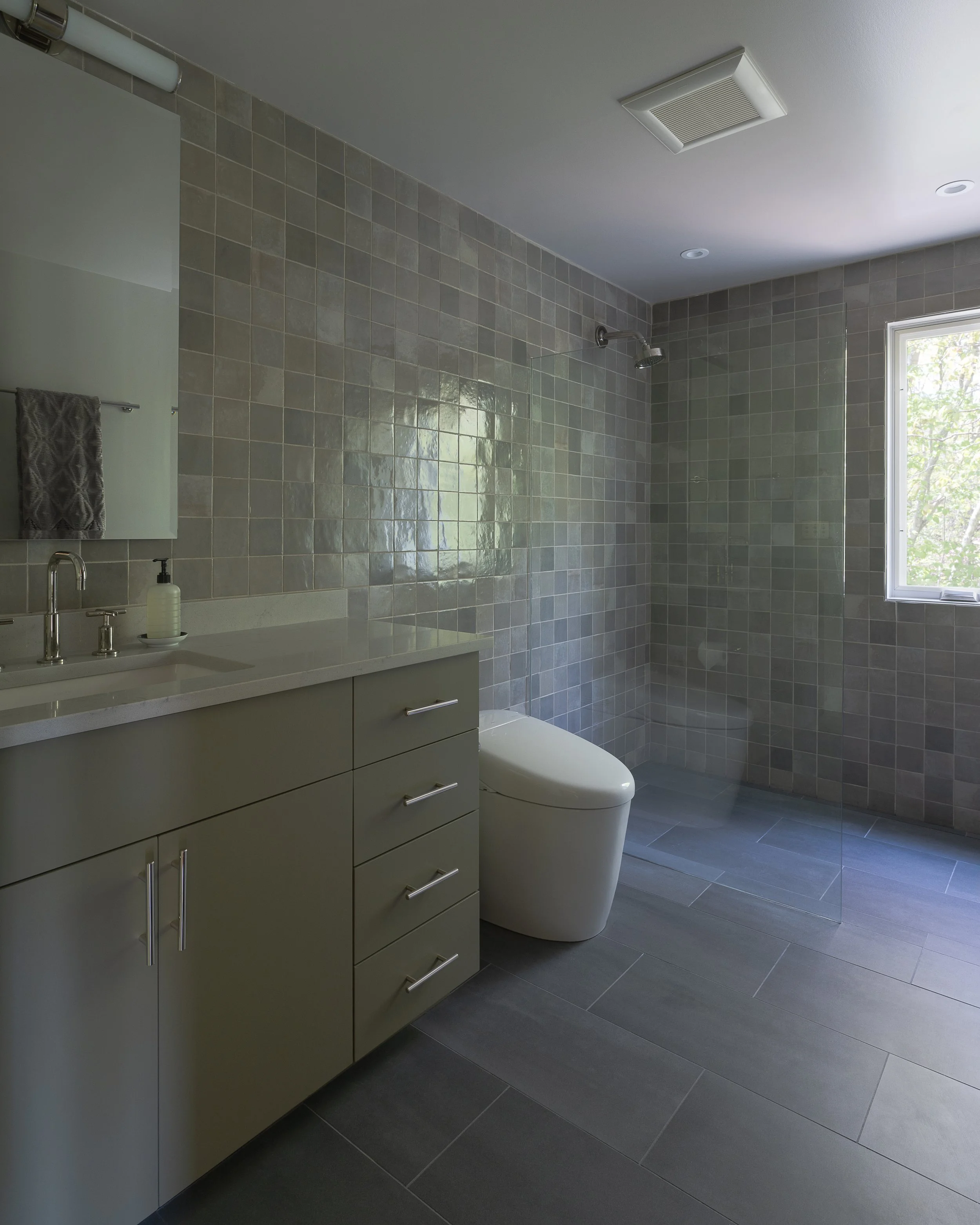Modern bathroom with gray tiled walls and floors, a white vanity with a mirror, a white toilet, a glass shower door, and a window providing natural light.