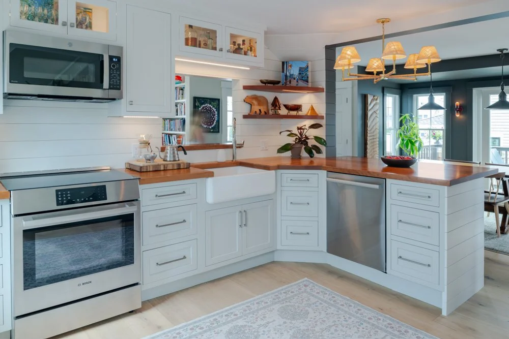 Modern white kitchen with wooden countertops, stainless steel appliances, open shelving with decorative items, and a chandelier over the island.