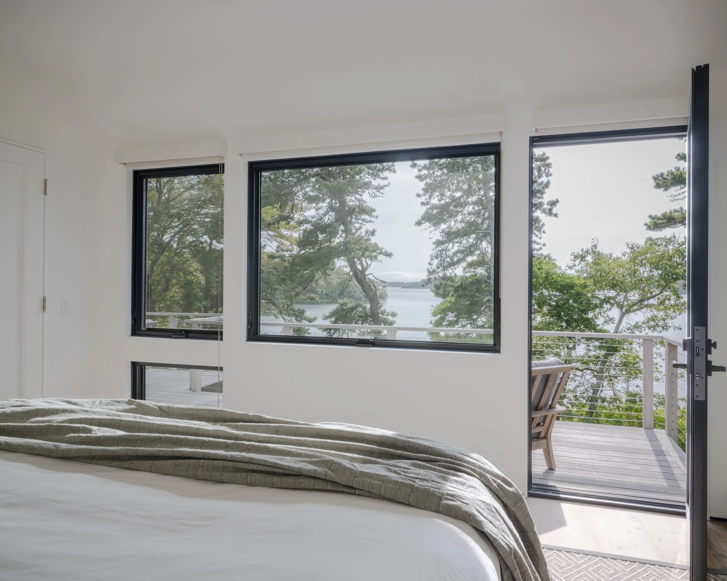 A bedroom with a bed and a balcony door open to an outdoor deck with trees and water in the background.