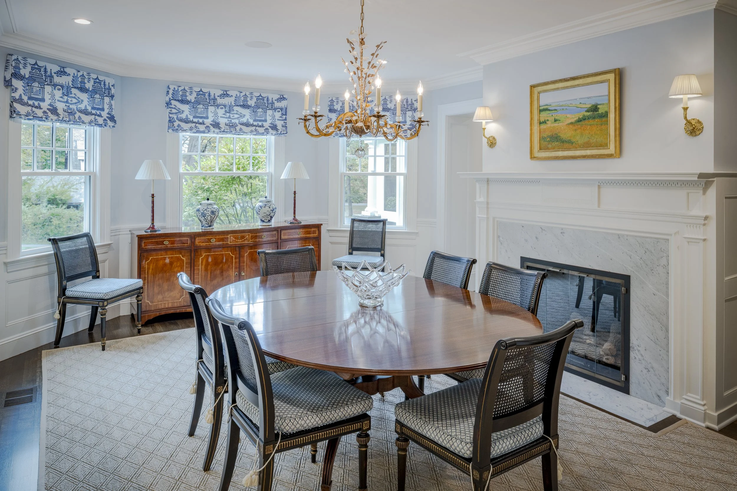Elegant dining room with a wooden oval table, eight upholstered chairs, a marble fireplace, a chandelier, and three windows with blue and white patterned valances, natural light, and decorative vases on a sideboard.