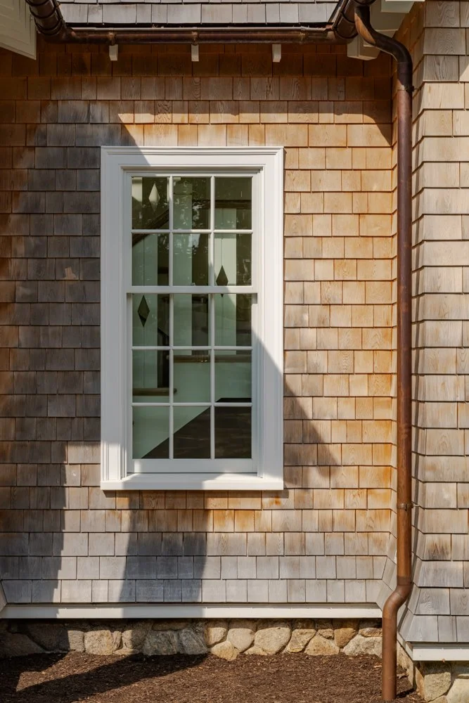 A house exterior with wooden shingle siding, a white-framed window with diamond-shaped glass accents, a metal gutter, and a stone foundation.