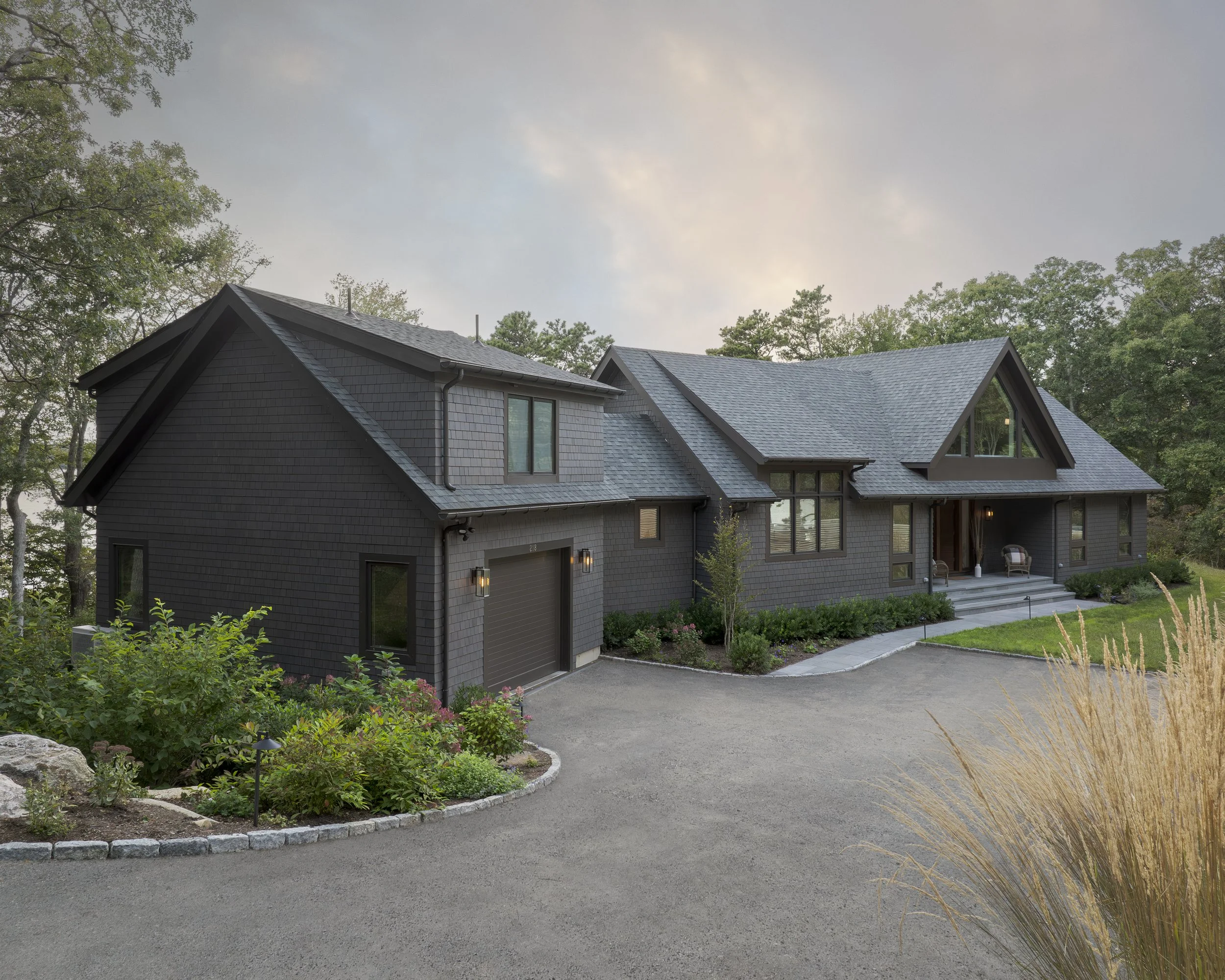 A modern, dark gray house with a steep roof and front porch, surrounded by a landscaped yard with bushes, flowers, and tall grasses, with trees in the background and a paved driveway.