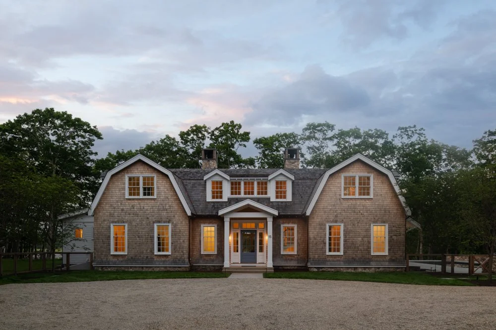 A large, rustic house with a shingled exterior, multiple dormer windows, and a central front door illuminated from inside. Surrounded by trees with a gravel driveway in front.