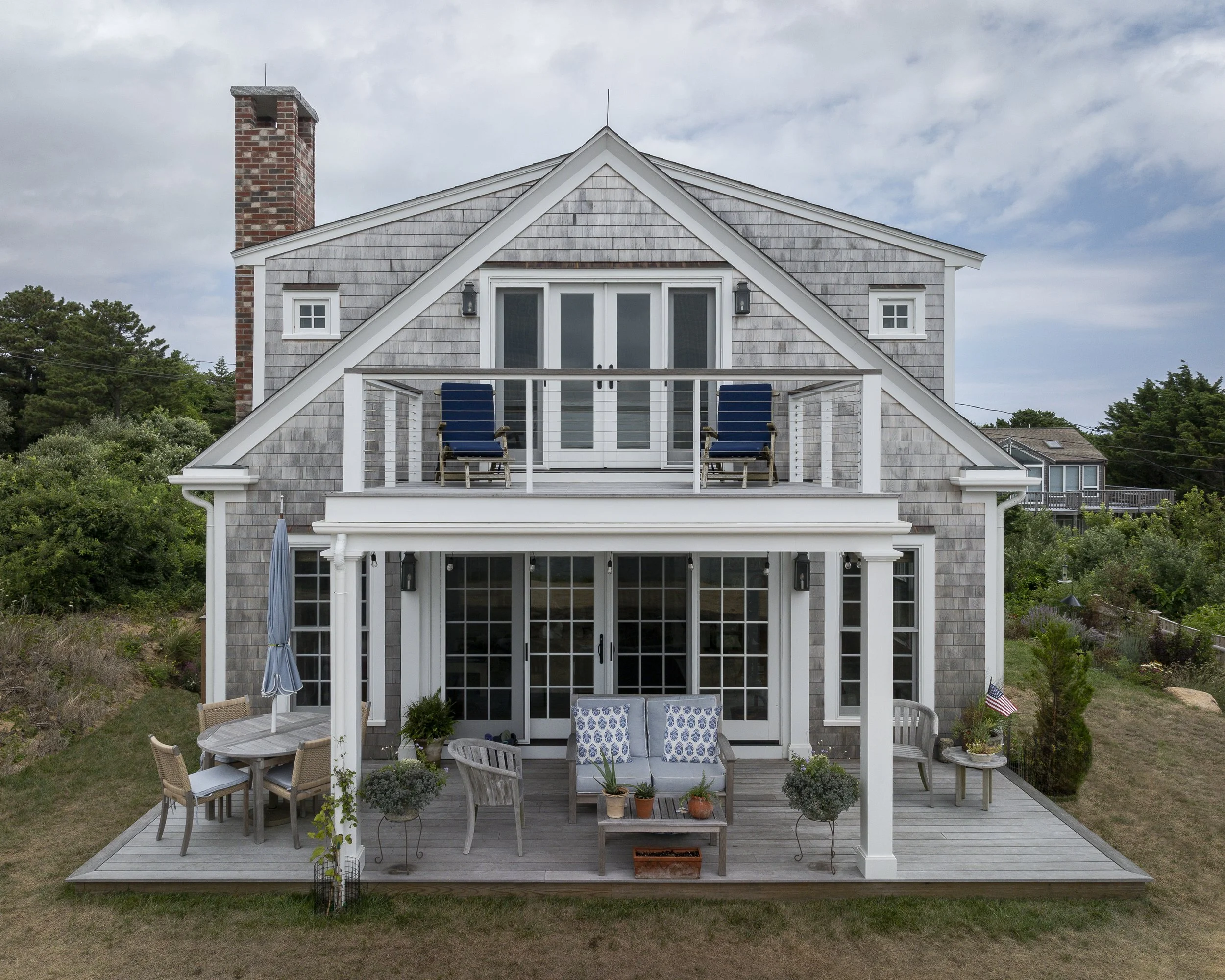 A two-story house with gray shingle siding, a chimney, and a wooden deck with outdoor furniture, including chairs, a table, and potted plants, under a partly cloudy sky.