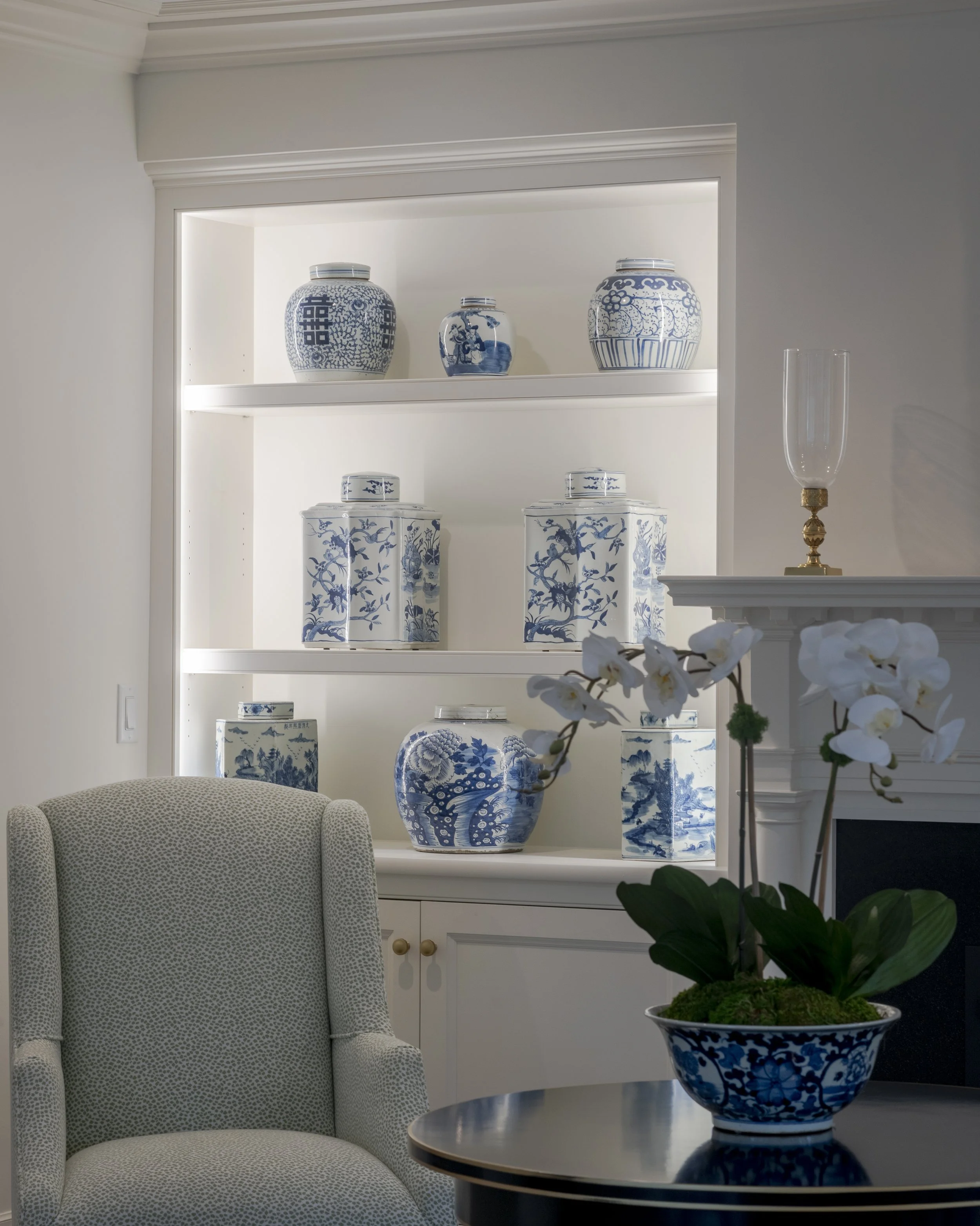 A white built-in display case with shelves holding blue and white ceramic jars and vases, with an orchid plant in a decorative blue and white bowl in the foreground, an upholstered armchair, and a fireplace with a glass hurricane lamp.