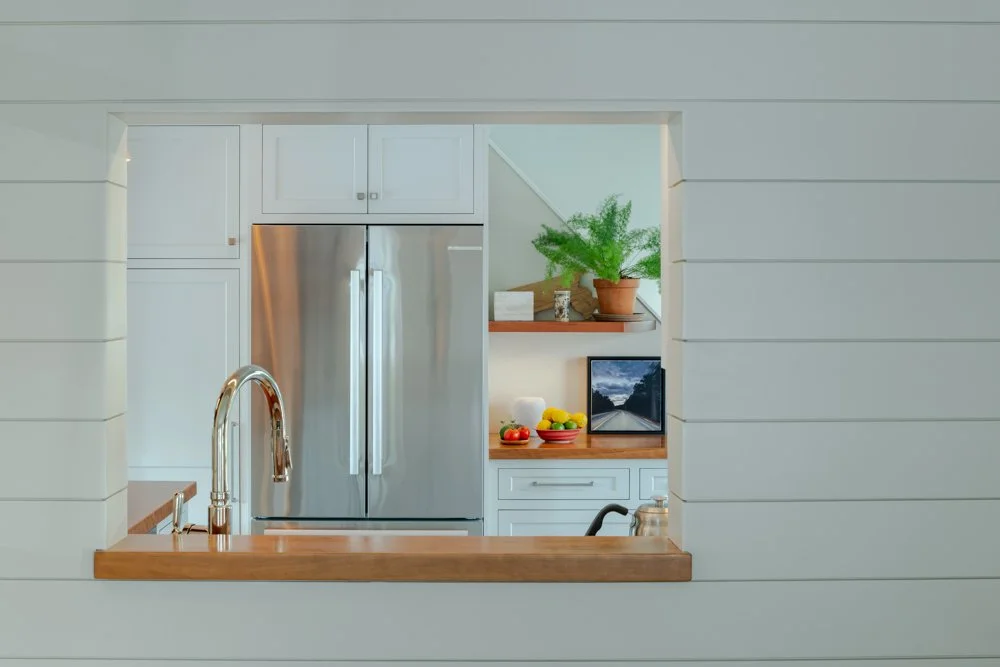 View of a modern kitchen through a square opening in a white wall, featuring a stainless steel refrigerator, wooden countertop, green plant, and a small electronic device.