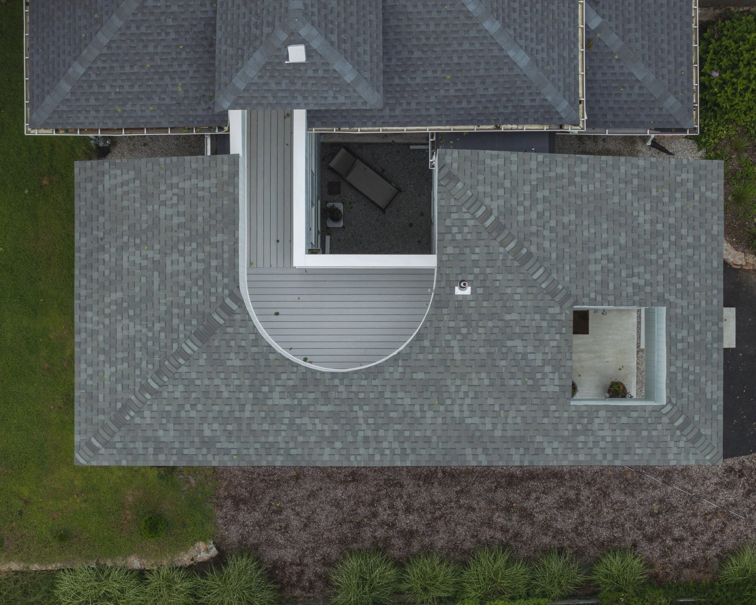 An aerial view of a house roof with gray shingles, including a small balcony with outdoor furniture and potted plants, surrounded by green grass, bushes, and a gravel path.