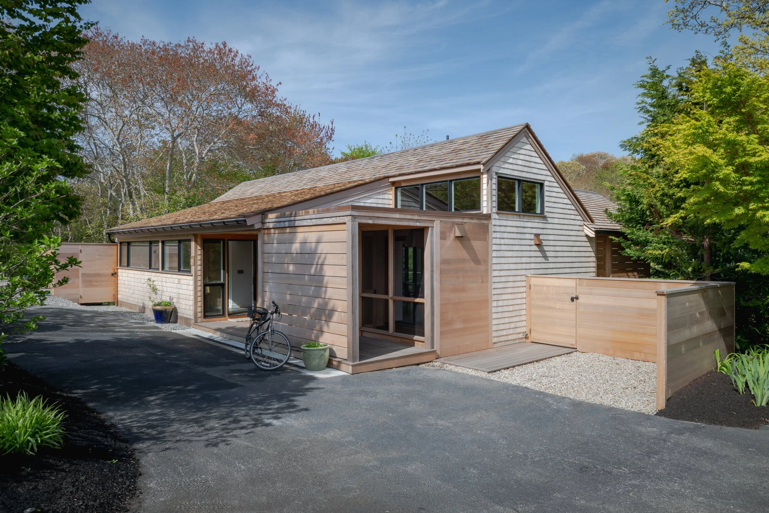 A modern house with a mix of wooden and shingle exterior, large windows, a small porch with a bike, and landscaped yard with trees and plants.