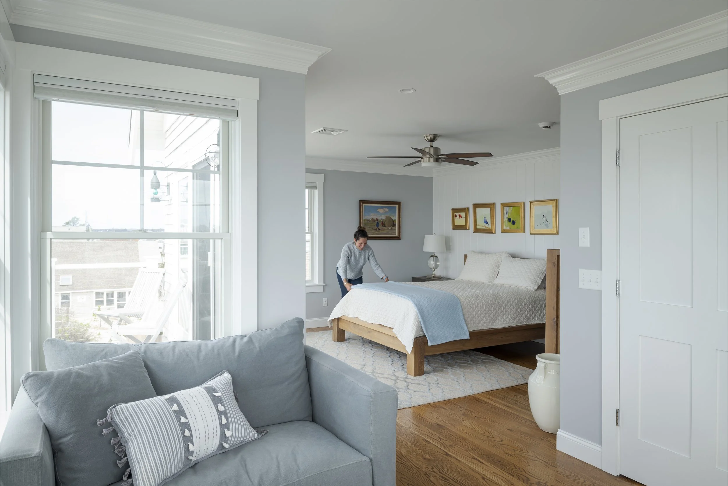 Interior of a bedroom with a woman making the bed, a ceiling fan, artwork on the wall, a small bedside table with a lamp, a large window with blinds, and a light-colored sofa in the foreground.