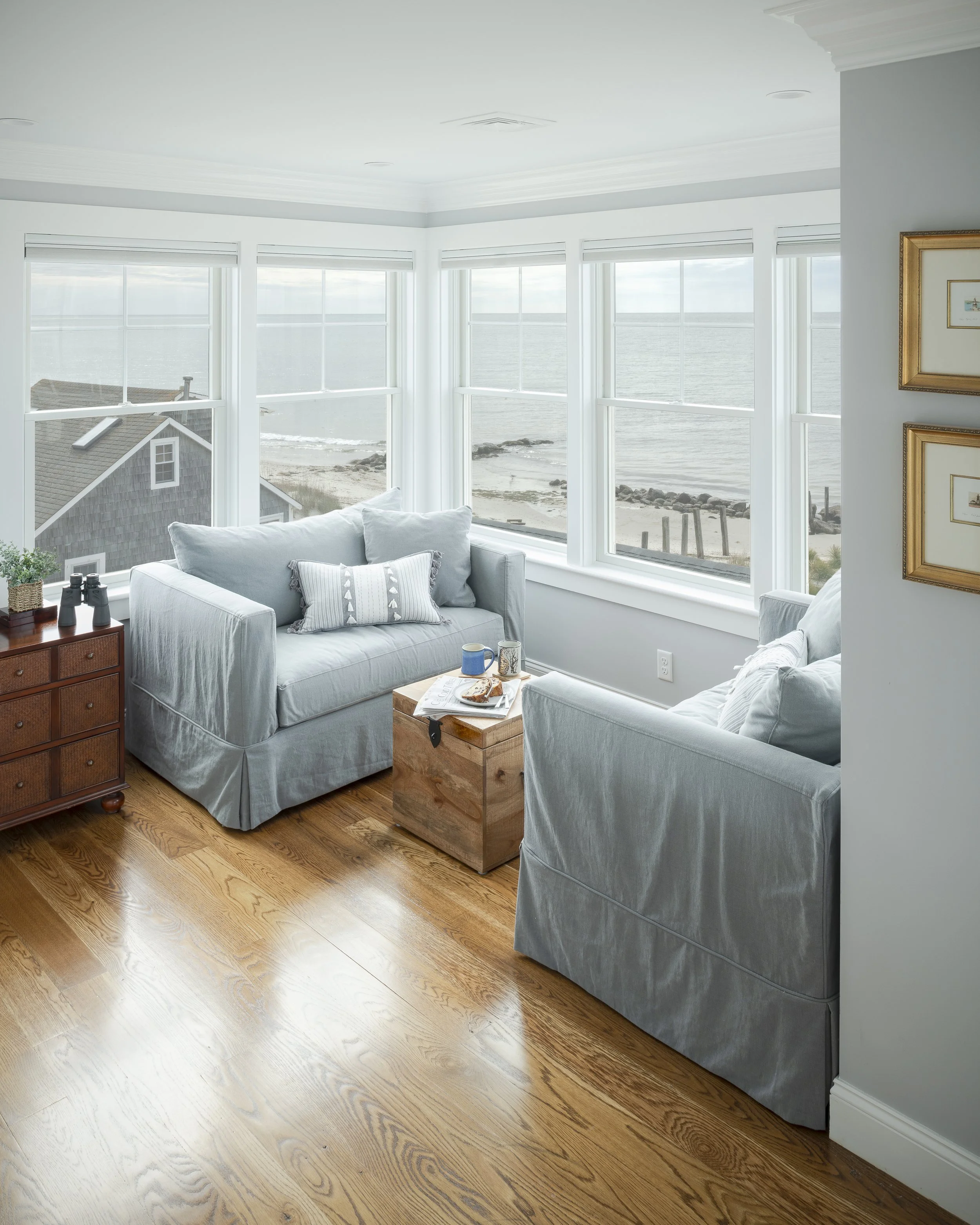 Living room with large windows overlooking a beach and ocean, featuring two light-colored sofas, a wooden chest used as a table with coffee cups and food, and framed pictures on the wall.
