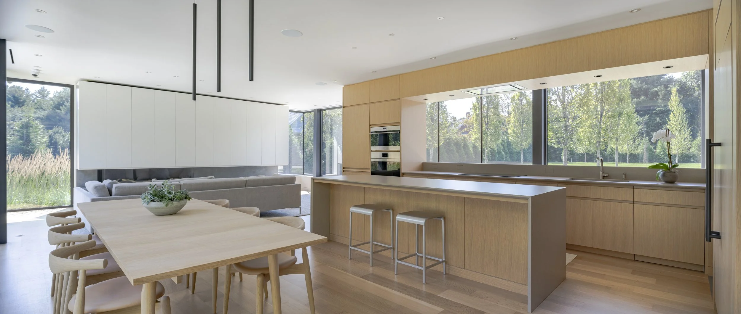Modern open-concept kitchen and dining area with light wood cabinetry, a large island, bar stools, and panoramic windows overlooking greenery.