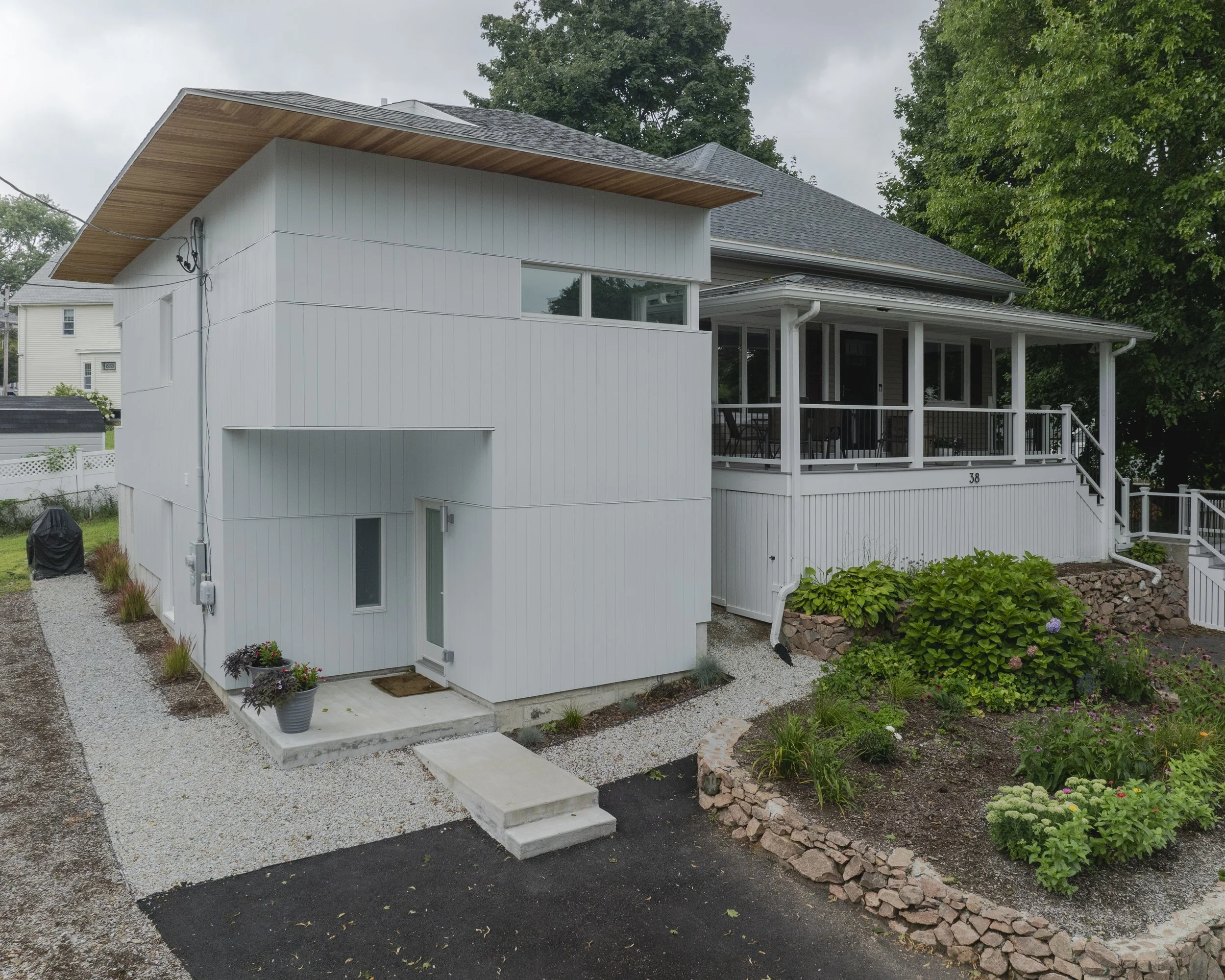 A modern two-story house with a white exterior, large front porch, and landscaped garden. The house features a sloped roof and a garden with various plants and flowers.