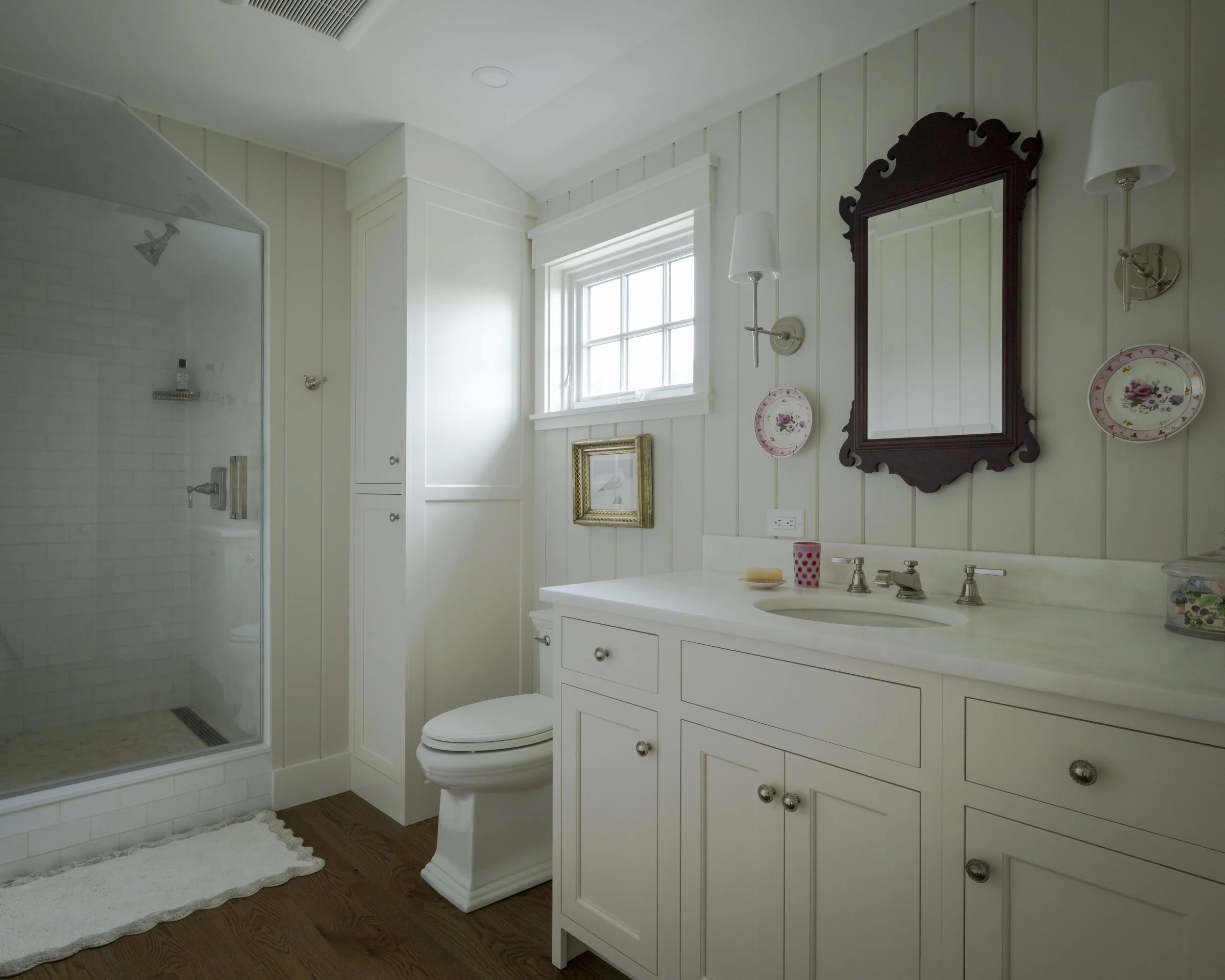 A bathroom with a white vanity, oval mirror, wall-mounted lights, a window, a shower with glass door, and wooden flooring.
