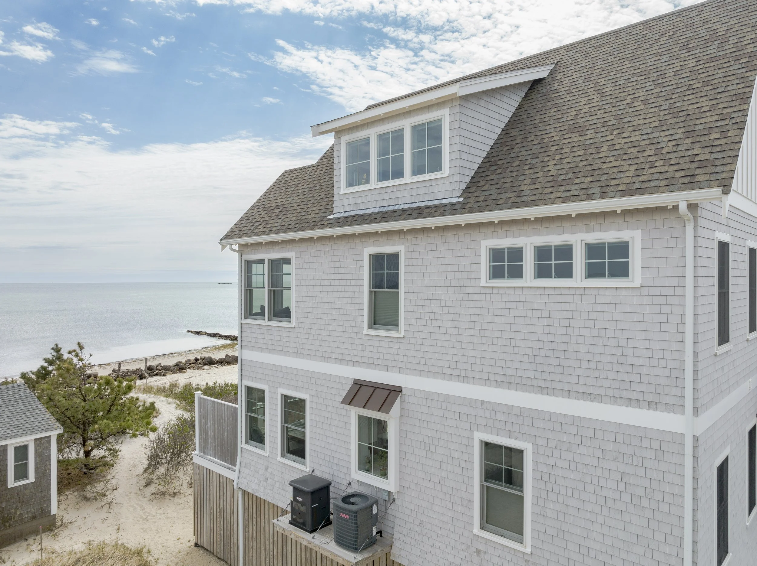 A multi-story beach house with gray shingles and white trim near the ocean, with sand and sparse vegetation in the foreground.