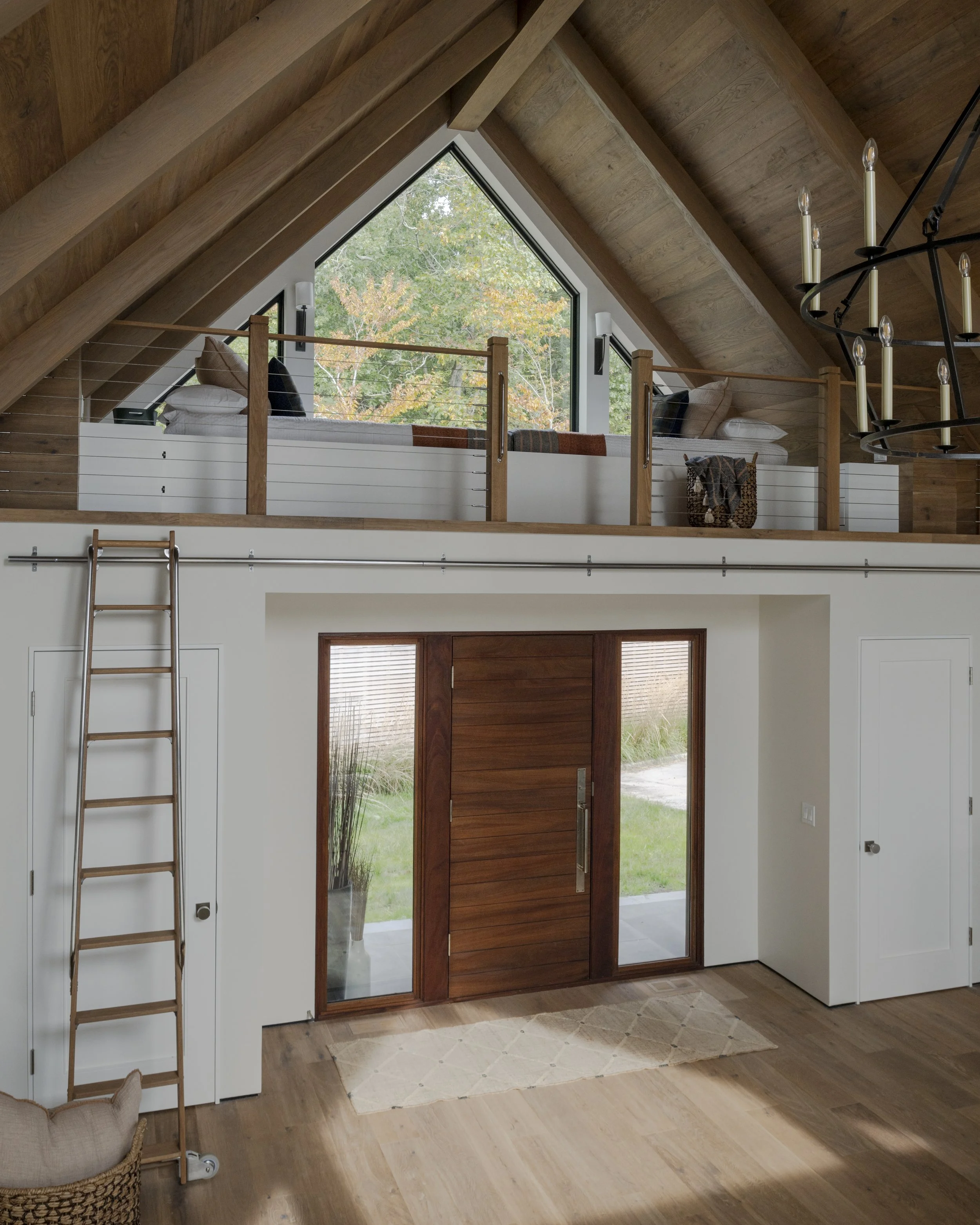 Interior view of a modern home with a high vaulted wooden ceiling, a large front door made of wood and glass, and a spacious entrance area with hardwood floors. A ladder leans against the wall near a closet, and a mezzanine with a window seating area