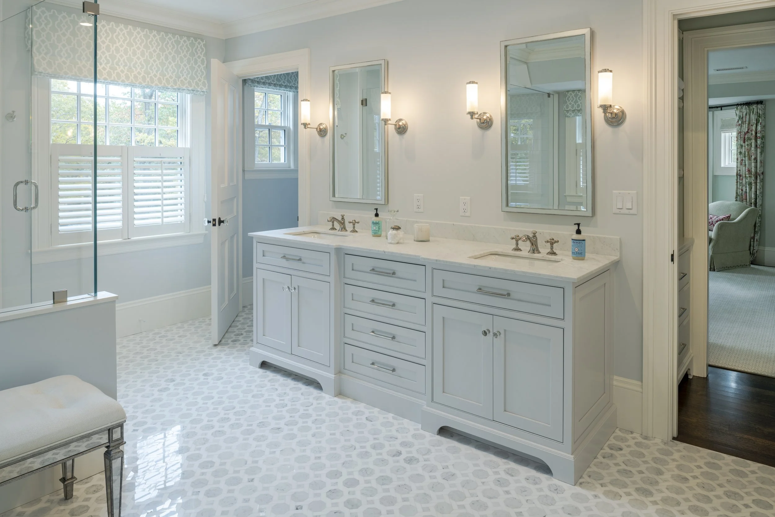 Bright bathroom with double vanity, white cabinets, marble countertop, and two mirrors. Light fixtures above mirrors, window with plantation shutters, and a glass-enclosed shower. Adjacent room visible through door.