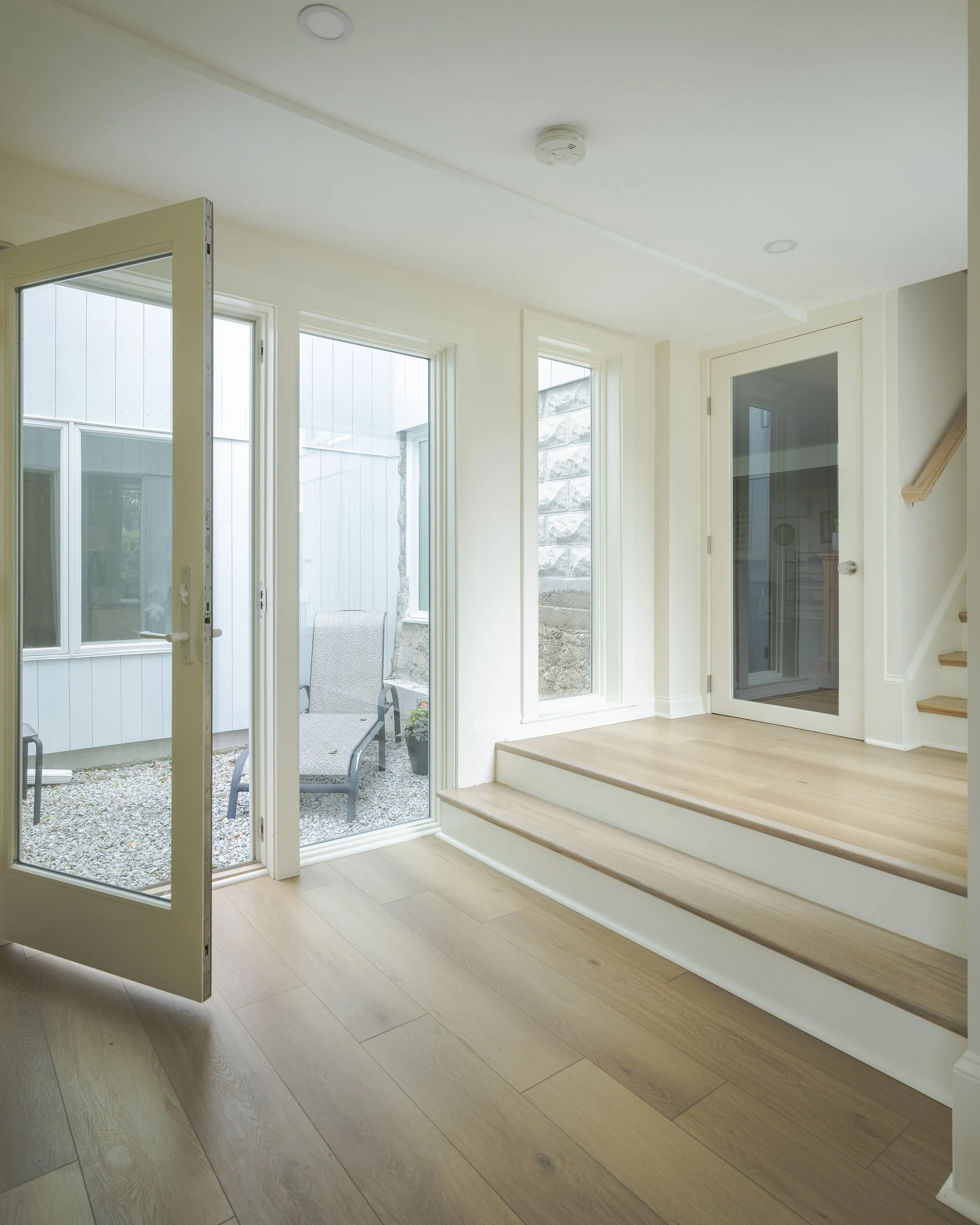 Interior view of a bright room with wooden flooring, a glass door leading outside, and a step up to another small wooden platform, with large windows and a staircase to the right.