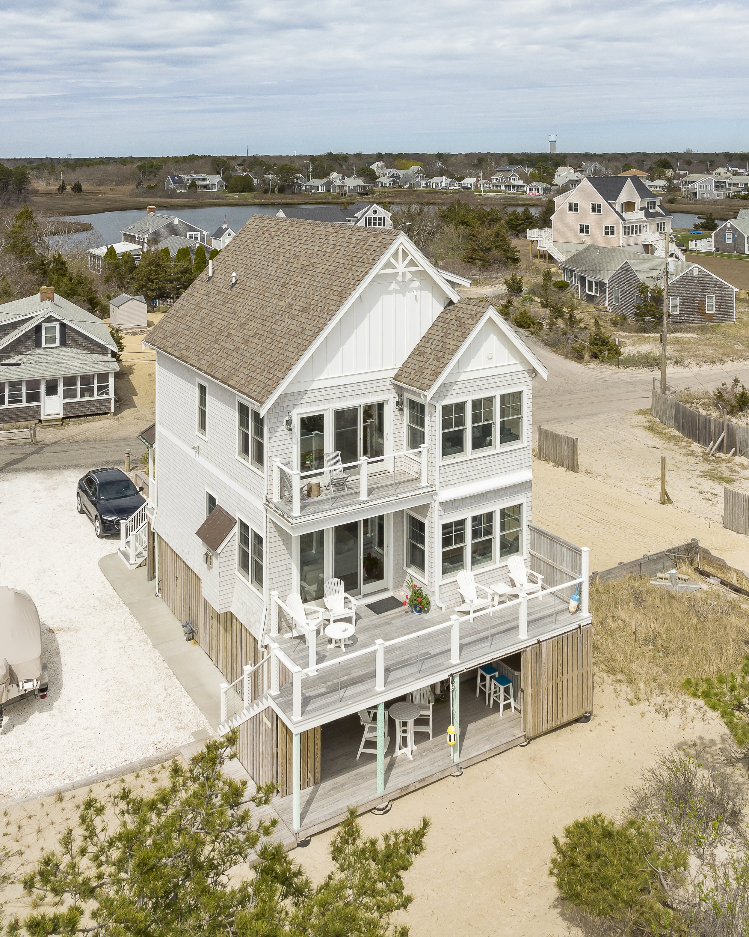 A three-story beach house with white exterior, large decks with seating, surrounded by sand and neighboring houses near a water body.