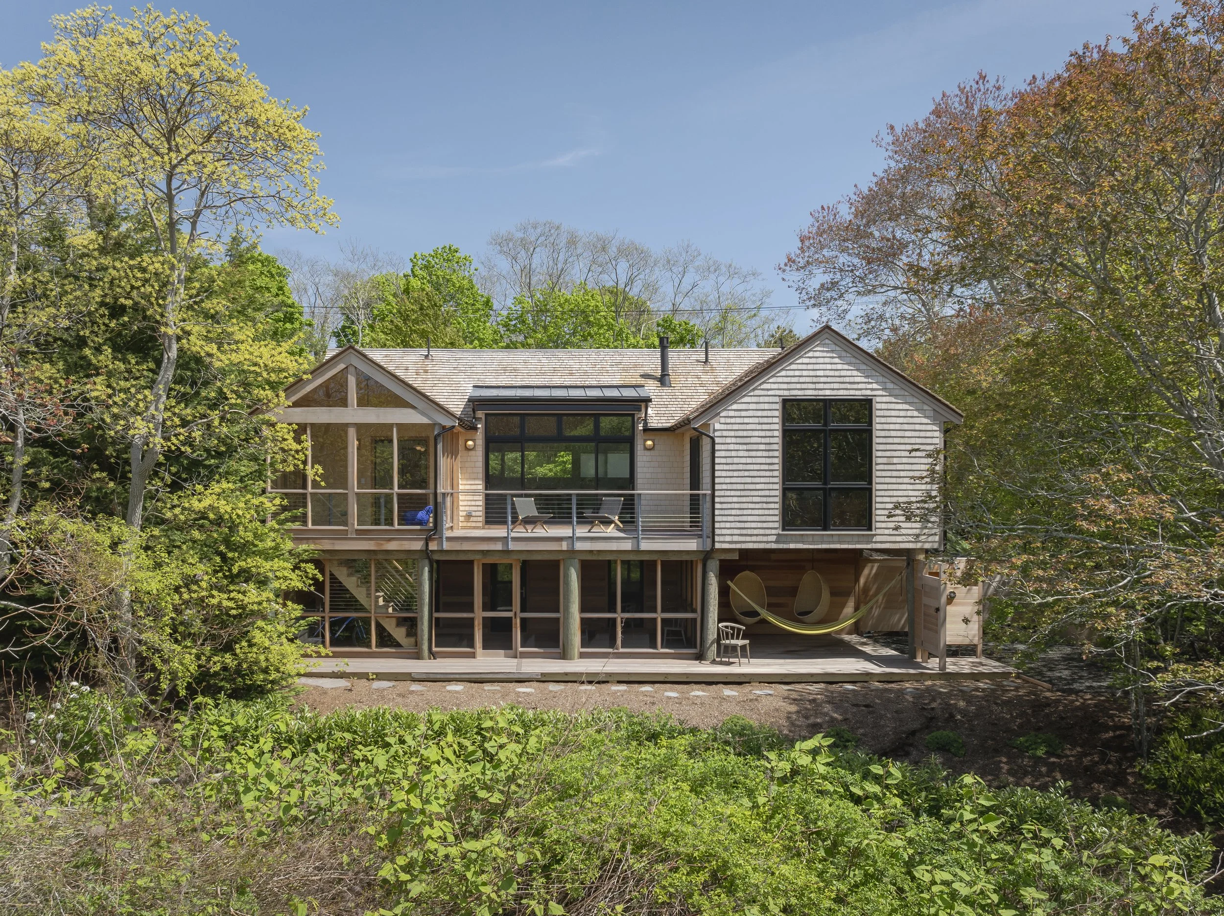 A two-story house with a screened porch and outdoor deck, surrounded by trees and greenery, under a clear blue sky.