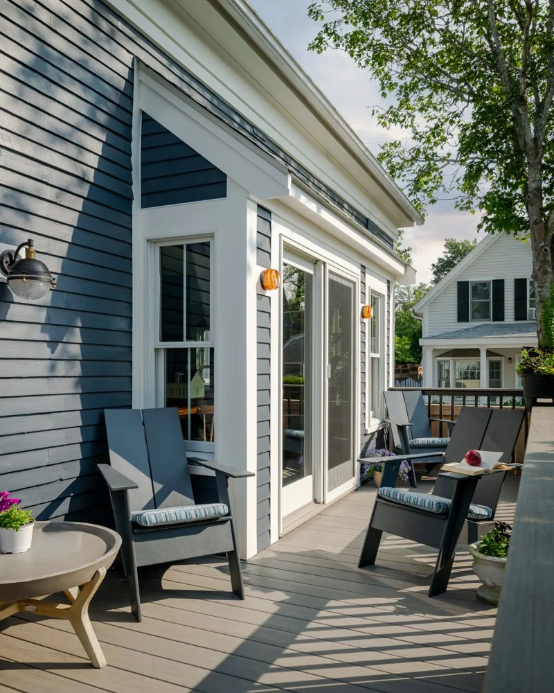 A backyard deck with blue siding, four chairs, small tables, potted plants, and a view of neighboring houses.