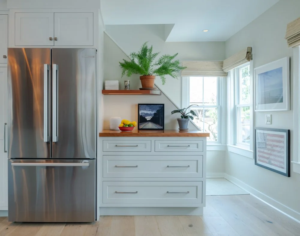 Modern kitchen with stainless steel refrigerator, white cabinetry, wooden countertop, potted plants, framed artwork, and large windows with beige blinds.