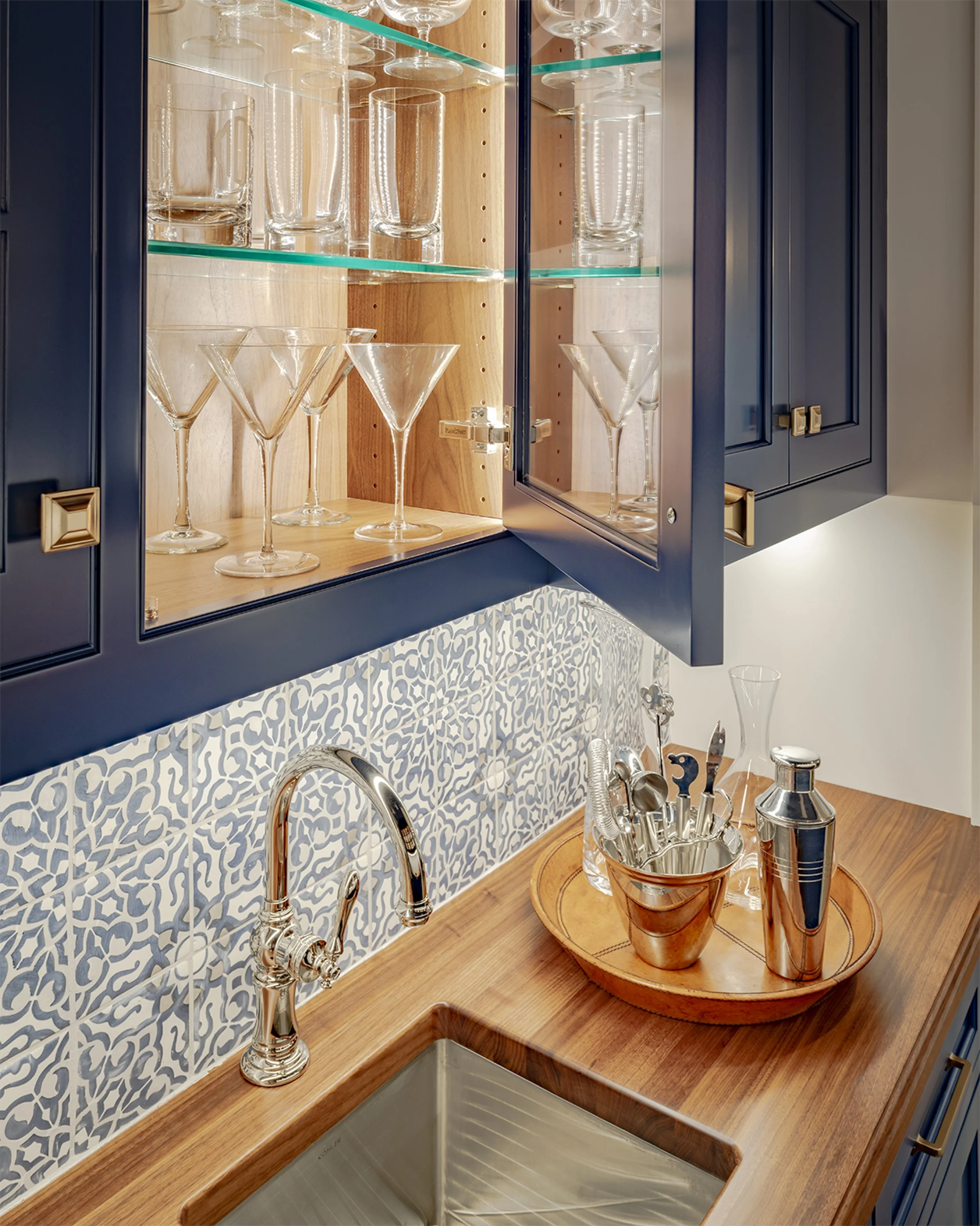Kitchen cabinet with glassware, wooden countertop with sink, and bar tools in a metal container.
