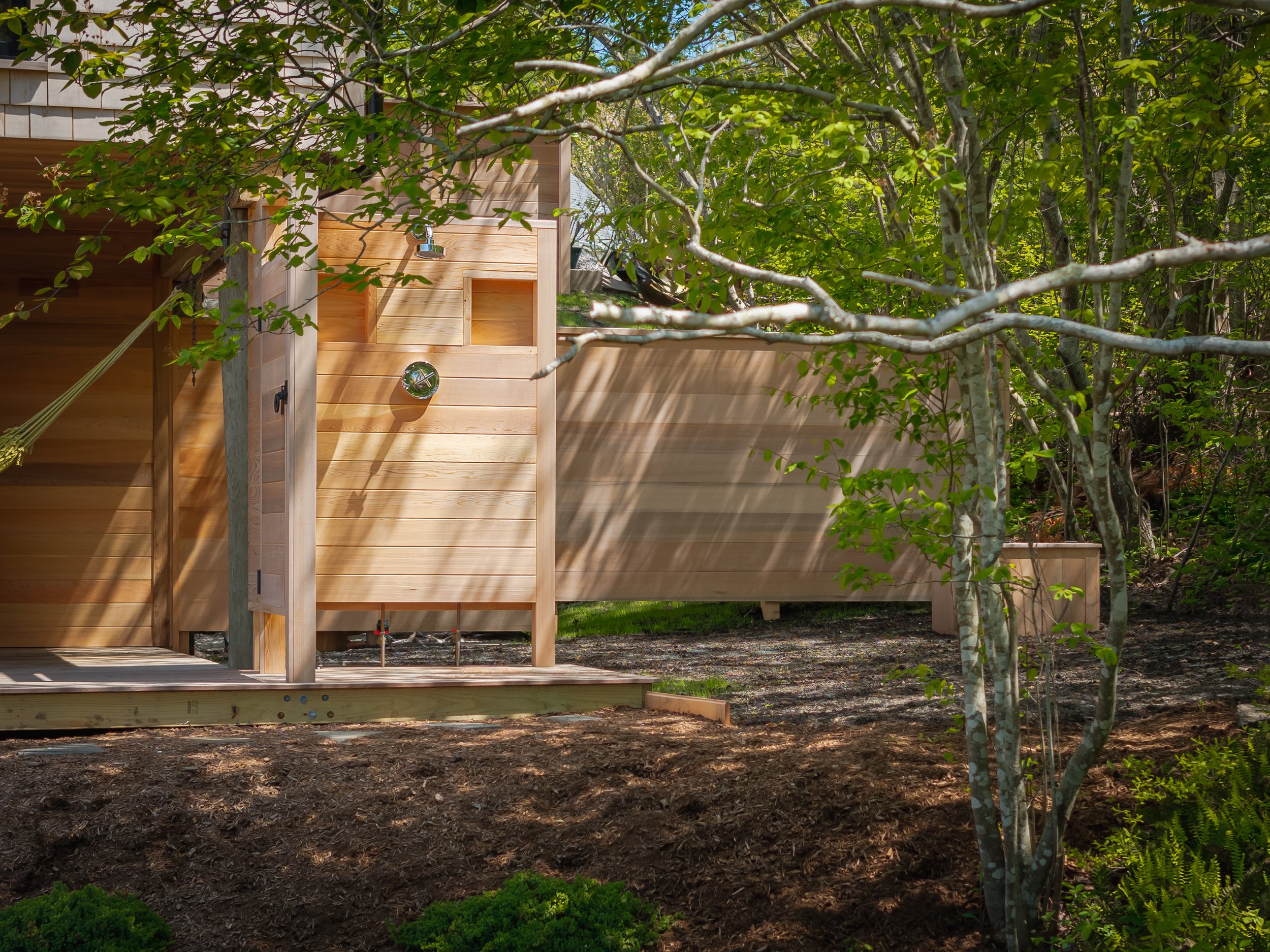 A wooden outdoor shower with a wall, set in a lush green garden with trees and shrubs.