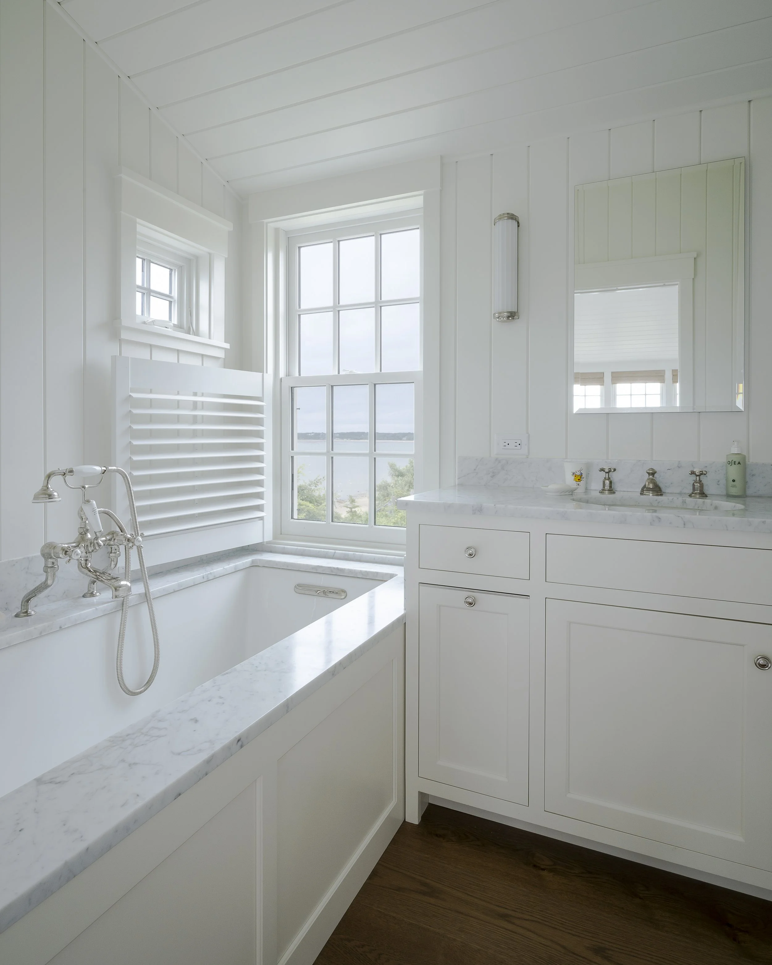 Bright bathroom with a white bathtub, marble countertop, and a window overlooking water and trees, with white cabinets and wooden floor.